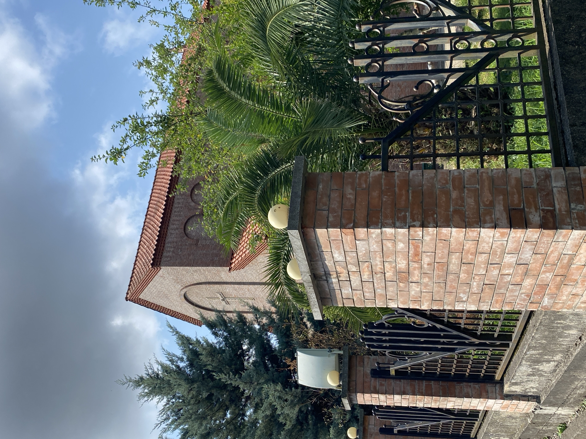 The image depicts a brick building with a red tiled roof, possibly a church, featuring arched windows and a cross visible on its facade. The building is partially obscured by lush green foliage, including a palm tree and a tall evergreen. In the foreground, a brick fence with decorative black metal gates runs along the front. The fence posts are topped with spherical decorative elements, and one post features a light-colored fixture, likely a security light. A concrete wall forms the base of the fence. The scene is set against a backdrop of a partly cloudy sky, with the overall environment suggesting a temperate climate. The presence of a palm tree and the architectural style hints at a location in Batumi, Georgia, a city known for its subtropical climate and diverse architecture. There are no people or active events captured in this still image.