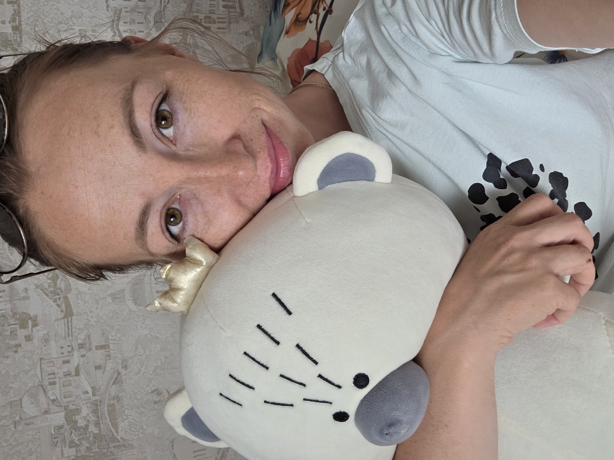 The image shows a woman lying on her side, possibly on a bed, and embracing a plush toy that resembles a bear. The woman is fair-skinned with light brown hair and hazel eyes. She wears glasses and has a light-colored shirt with black spots on it. The plush toy is predominantly white with grey accents on the ears and nose. It has black embroidered dashes around the nose and a small golden crown on its head. The background appears to be a patterned wall or bedsheet. The image is taken in a close-up, intimate setting. The location of the scene is unspecified.