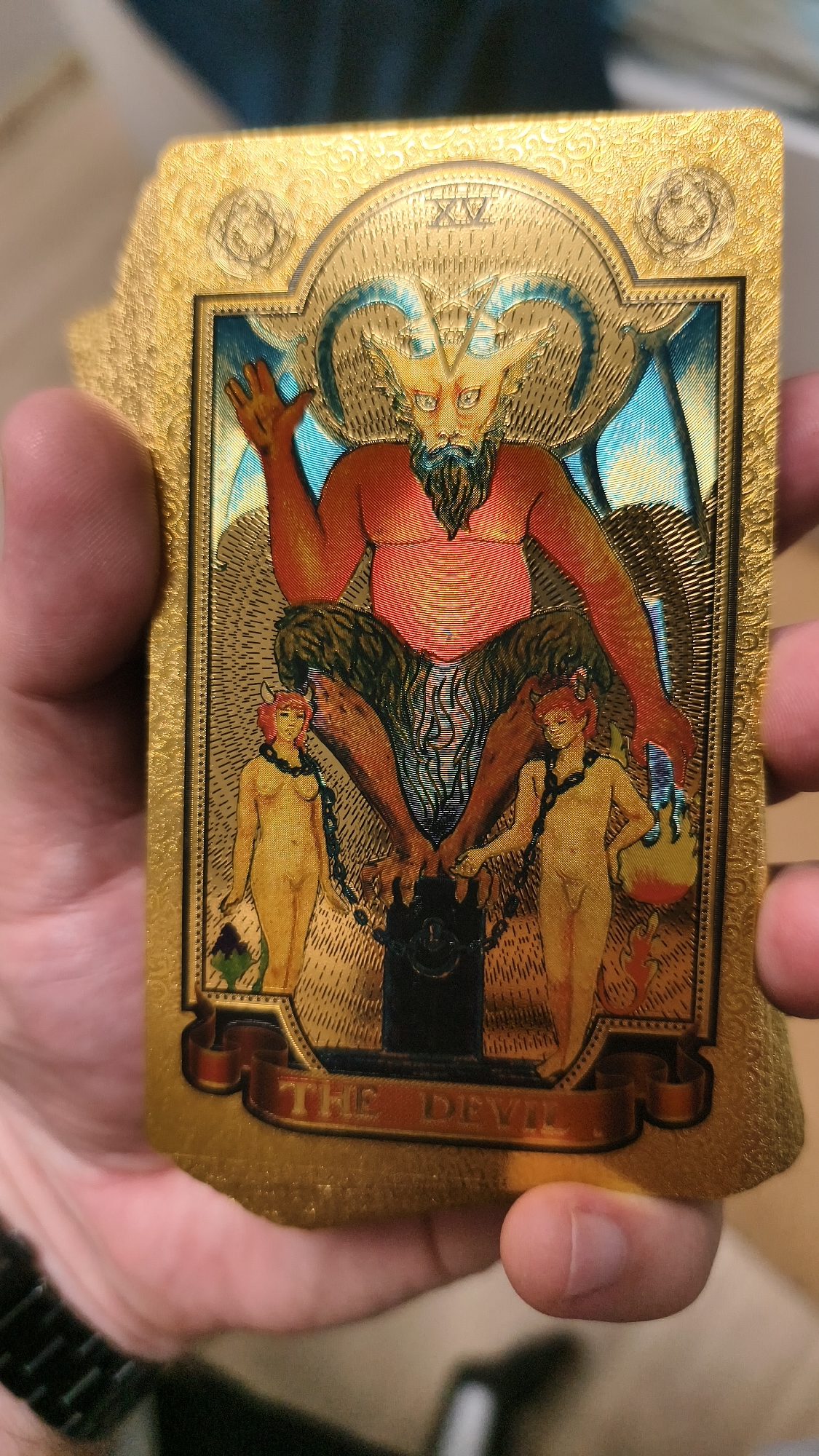 A gold-embossed tarot card, "The Devil," is held in a person's hand. The card depicts a seated, demonic figure with red skin, large horns, and a beard, flanked by two naked figures with tails. The background is textured and golden. The Roman numeral "XV" is visible at the top, indicating the card's number. The scene takes place in an indoor environment, with a blurred background suggesting an out-of-focus room.