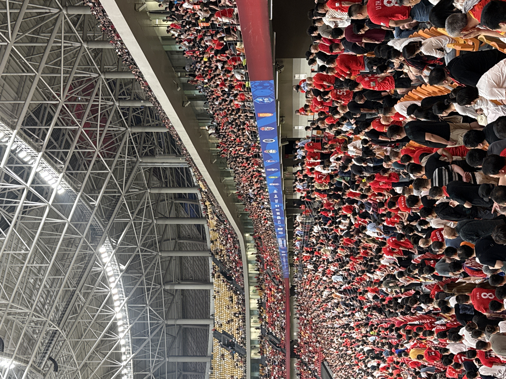 A tightly packed crowd of people is situated in an indoor stadium. The majority of the attendees are wearing red shirts. Overhead, a complex framework of metal beams and structures supports the roof of the stadium. A banner with different colored emblems spans the side of the frame above the crowd. The location is possibly within the city of Longxing, China.