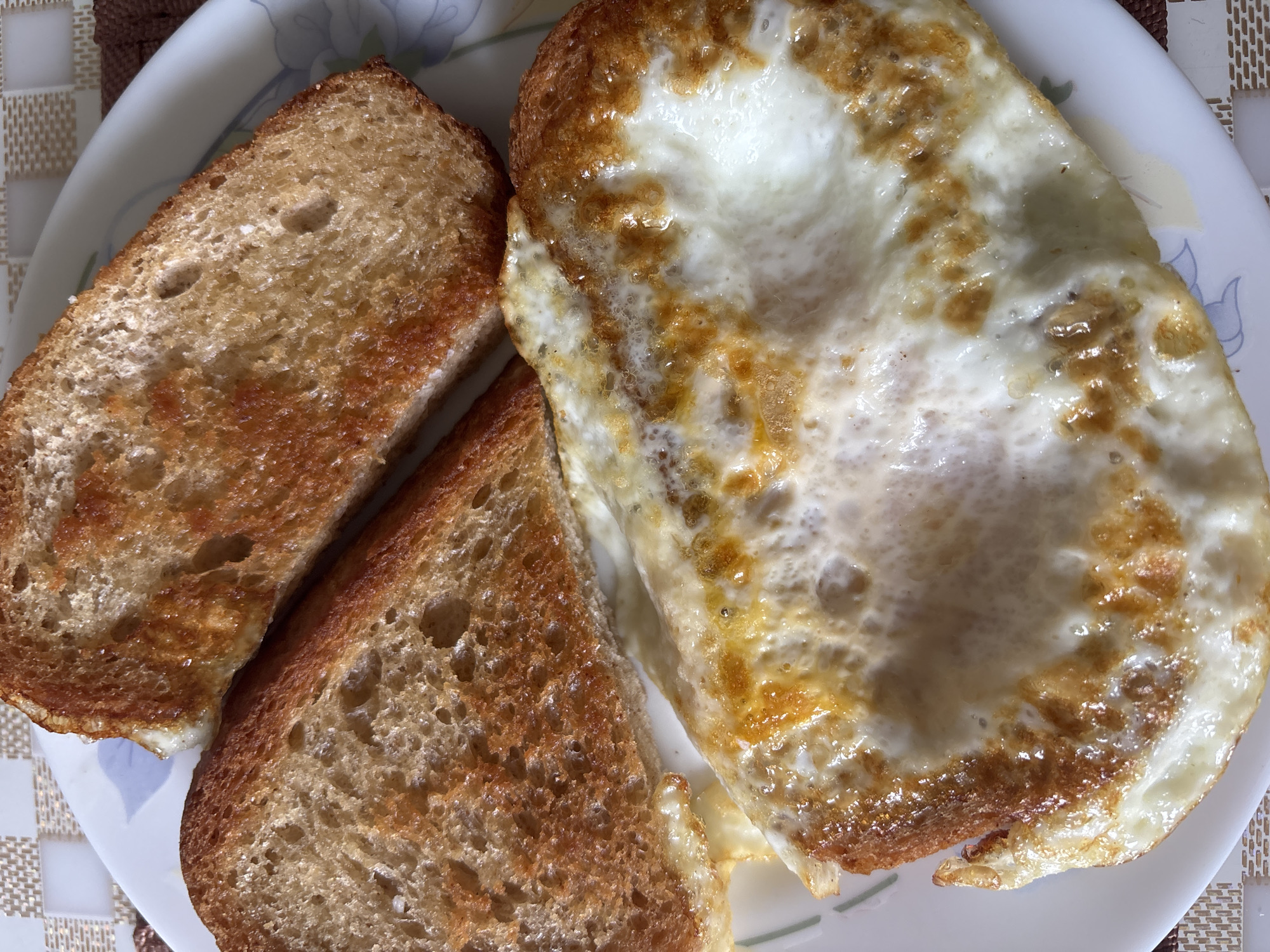 A close-up, overhead shot shows a plate of food with two slices of toasted bread and a fried egg. The toast is golden brown with visible pores and a slightly uneven texture. The fried egg is cooked sunny-side up, with a firm white and a bright yellow yolk that appears to be intact. The egg has slightly crispy edges and some browned bits from cooking. The food is placed on a white plate with a subtle floral pattern along the rim. The background shows a checkered placemat in light brown and beige tones. The lighting suggests it is daytime, with natural light casting soft shadows. The image was taken on a table in a residential setting, possibly a kitchen or dining room. There is no indication of the city of Bila Tserkva, Ukraine.