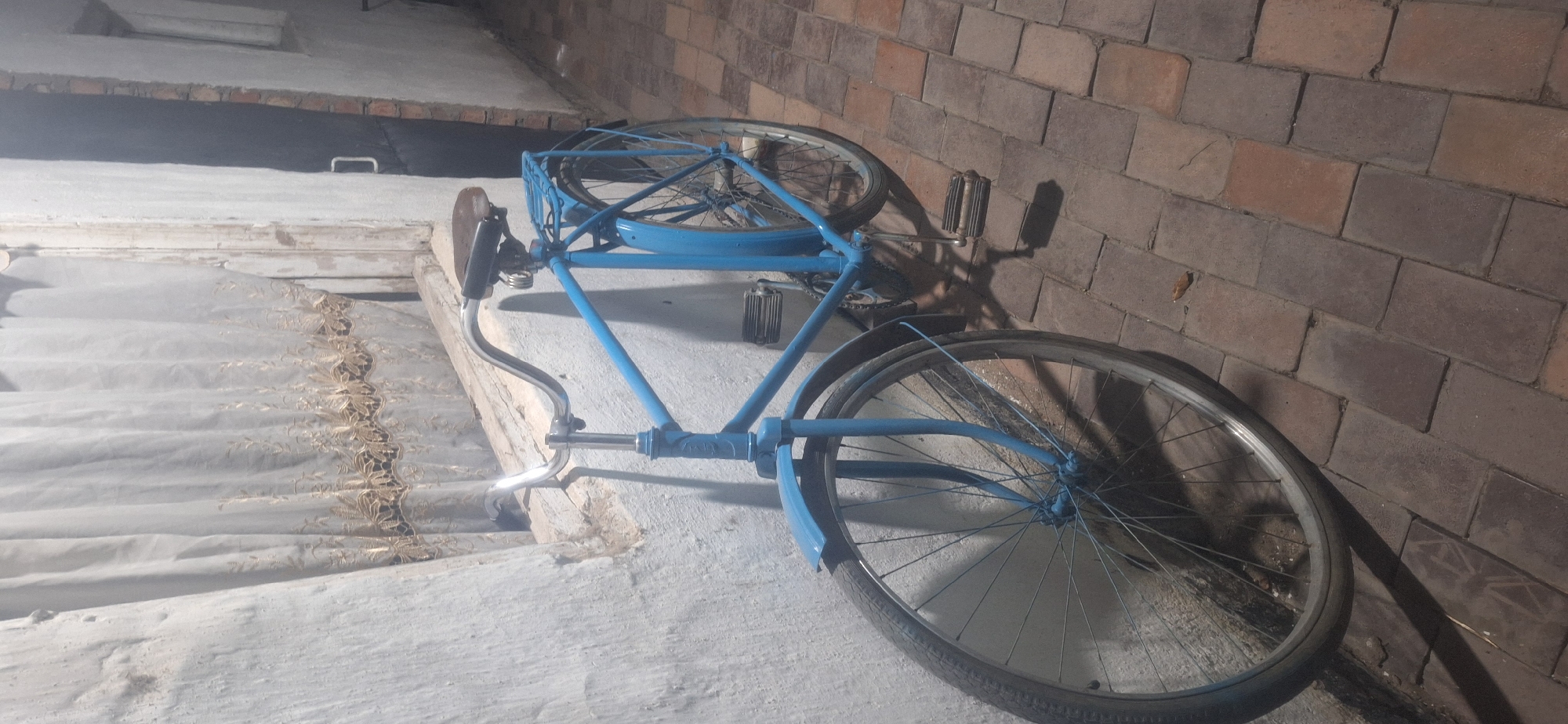 A vintage blue bicycle is parked against a textured, white wall, its handlebars curved and a brown saddle visible. The bicycle has a metal rack over the rear wheel, and both wheels have thin spokes. The scene is illuminated by artificial light, casting shadows on the brick-paved ground. The wall appears to be part of an outdoor structure, with a glimpse of a window covered by a sheer curtain with embroidered detailing visible to the left. To the right of the bicycle, a dark doorway or opening is visible, leading into a darker area. The environment suggests an exterior setting in Novopokrovka, Kyrgyzstan, possibly a courtyard or alleyway. No people are present in the image.