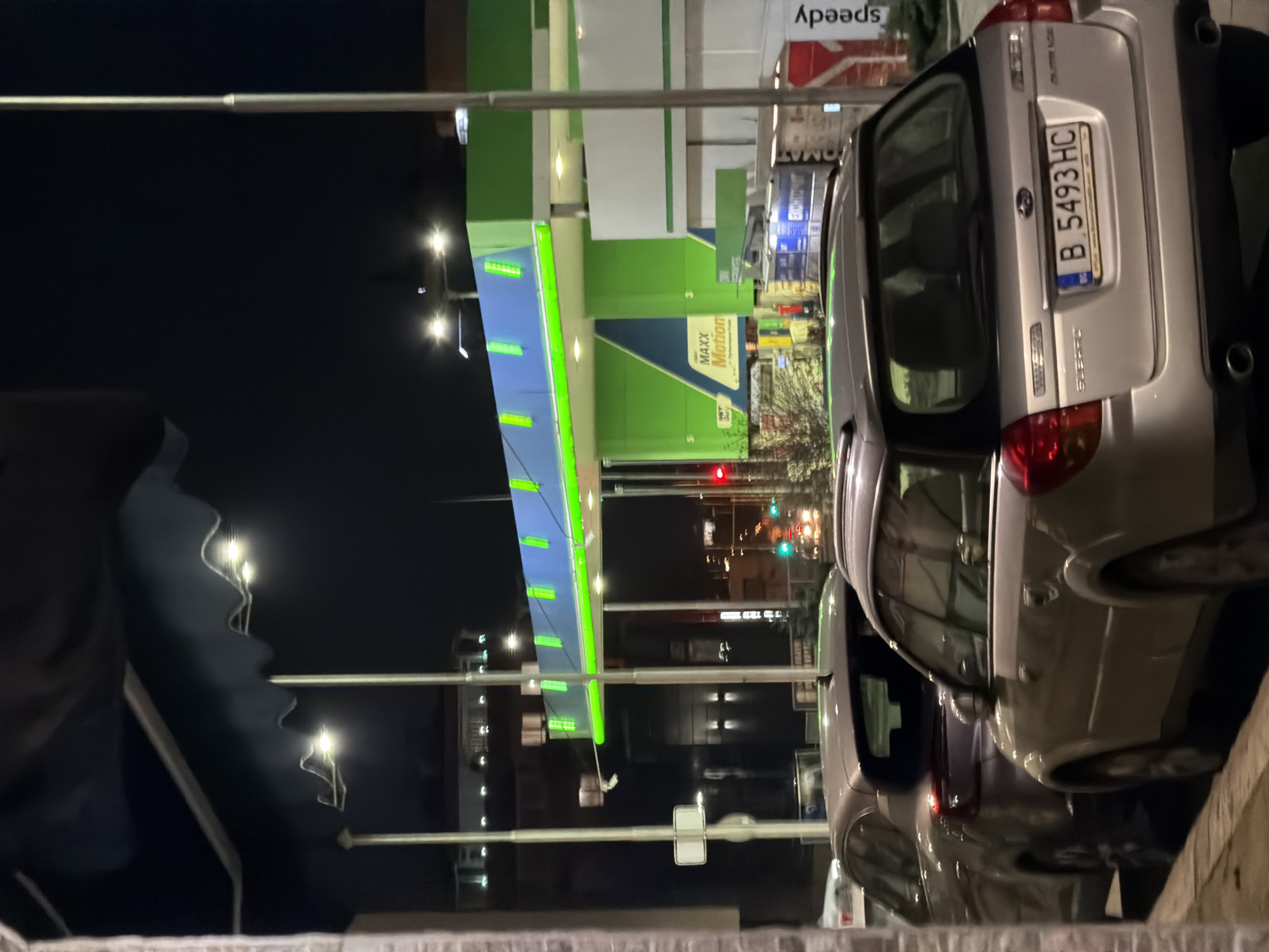 The image depicts a gas station at night in Varna, Bulgaria. The gas station is green and white, with "Maxx Motion" branding visible. Several streetlights illuminate the area. Two cars are parked nearby. The car closest to the camera has a license plate reading "B 5493 HC". Traffic lights are visible in the distance. The image appears to have been taken from a low angle, possibly through a fence or barrier. The foreground is slightly obscured.