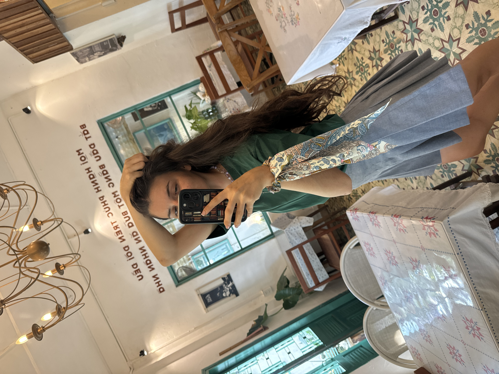 A young woman is taking a mirror selfie in a brightly lit cafe. She has long, dark brown hair, which she is tucking behind her ear with her right hand. She is wearing a green t-shirt and a grey pleated skirt. A colorful scarf is tied around her left wrist. Her phone, which she is using to take the photo, is black with a patterned case.
The cafe has a white ceiling with a large, ornate, multi-arm chandelier hanging from it. The walls are white, with Vietnamese text printed in red above a turquoise-framed window. There are several tables covered with white tablecloths patterned with pink and blue designs, and wooden chairs are arranged around them. The floor is tiled with a colorful, geometric pattern. To the left of the woman, a large plant with broad leaves sits in a pot. The overall ambiance suggests a trendy cafe in a Vietnamese city, possibly Phu Thuy Ward, Vietnam, given the presence of Vietnamese text.