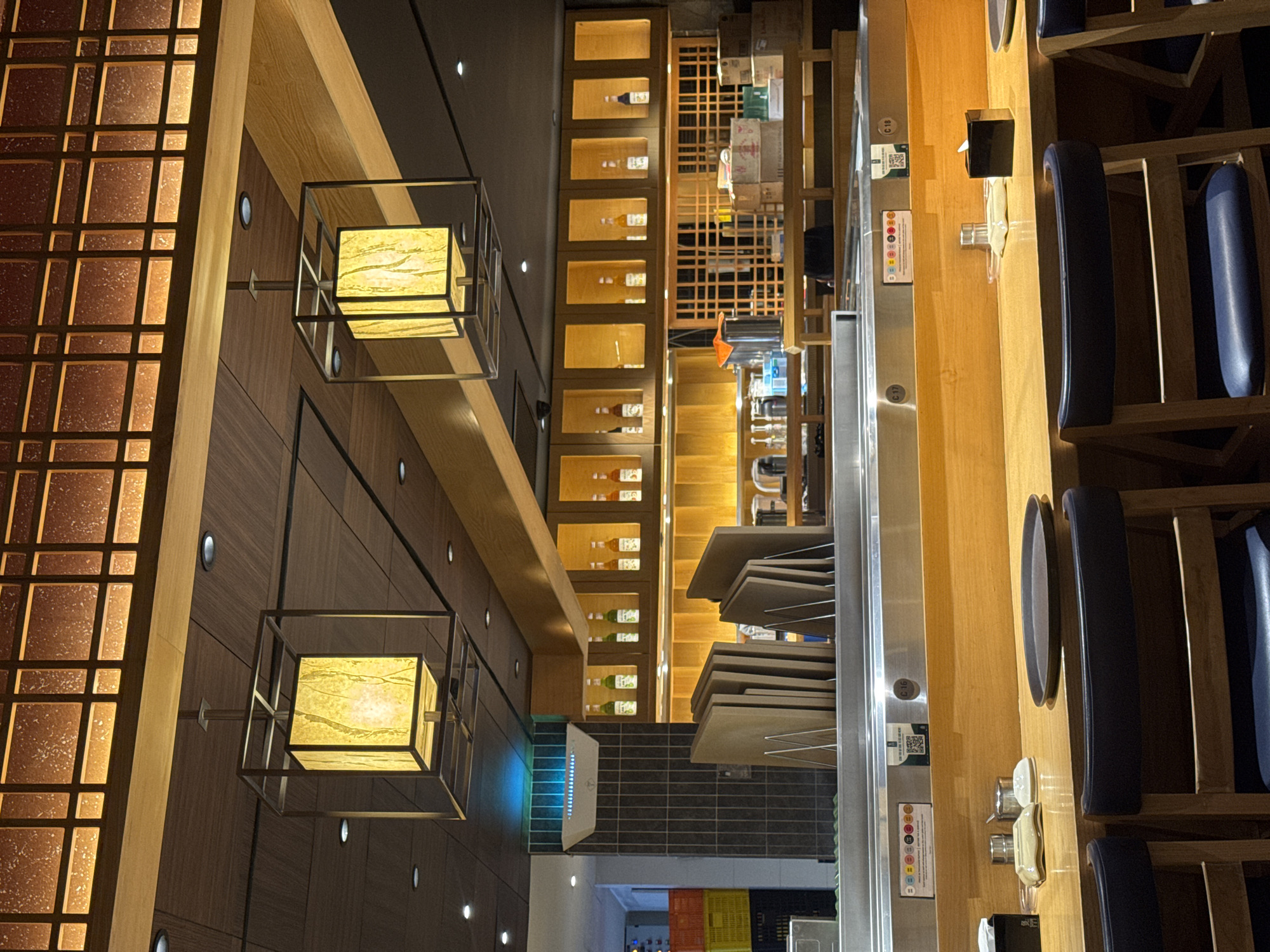 This image captures the warmly lit interior of what appears to be a modern, Asian-inspired restaurant or eatery in Kuta Selatan, Indonesia. The setting suggests an evening or night atmosphere, with artificial lighting illuminating the entire space.
On the left side of the frame, a decorative wall features dark wood paneling alongside a striking vertical screen with an intricate grid pattern, allowing light to diffuse through its squares. Suspended from the wall are two square-shaped lanterns with glowing, textured yellowish panels, adding to the ambient light.
Towards the center and background, a long series of recessed shelves, also made of light wood, display various bottles, likely beverages, each individually illuminated. Beyond these shelves, a glimpse of what might be a kitchen or service area is visible, with stacked trays and other restaurant equipment. Below the display shelves, a tiled section is visible, and a white air purification unit or similar device glows with blue light.
The right side of the image focuses on a long, wooden counter area, possibly a sushi bar or a casual dining counter. Several tall, wooden bar stools with dark blue upholstered seats are positioned along this counter. The counter surface features a metal strip, indicating a conveyor belt system or a service channel. On the wooden counter, various small items are placed, including condiment containers, small dishes, and a black holder for napkins or utensils.
Visible text includes "C 18" and "C 17" on the metal strip, likely seat or table numbers. Next to "C 18," a QR code is clearly visible, possibly for ordering or payment. There is also a strip with a series of colorful circles and symbols, which could represent dietary information or a rating system. No people are actively dining, but the setup suggests it is ready for patrons.