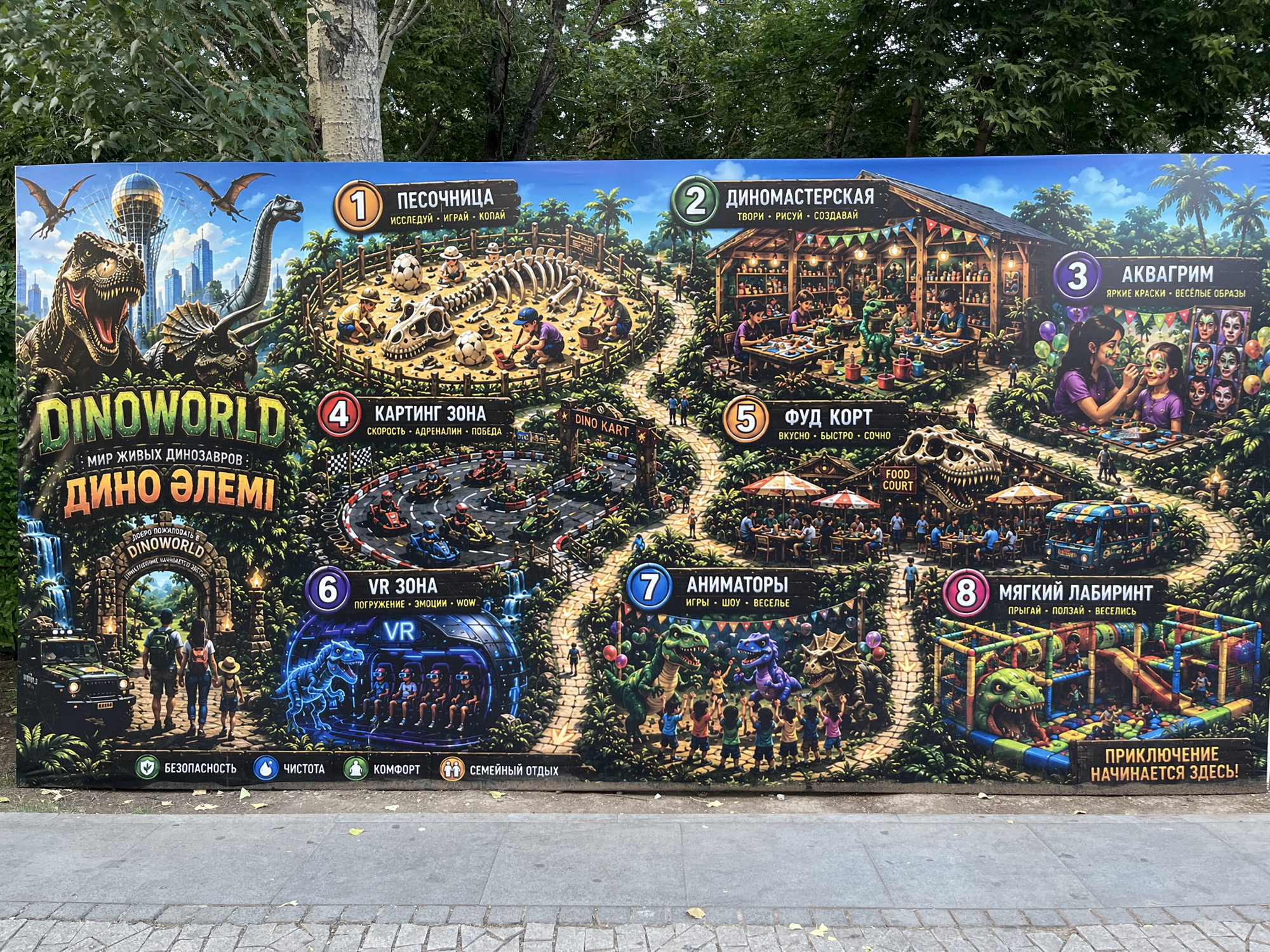 This is a photograph of a large, colorful mural advertising a dinosaur-themed amusement park called "DinoWorld" located in Astana, Kazakhstan. The mural depicts various attractions within the park, including a sandbox, a craft area, an aquarium, a karting track, a food court, VR experiences, animators, and a soft play area. The mural is positioned outdoors, with a park setting and trees visible in the background. The lower portion of the mural features icons and text related to safety, cleanliness, comfort, and family entertainment. The mural is mounted on a wall or fence and is in good condition, with no visible damage.