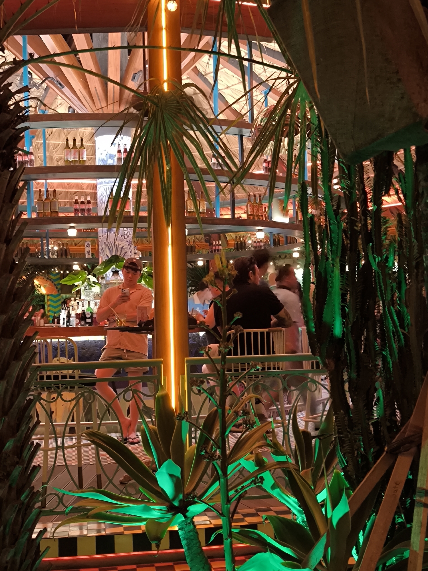 The image depicts a bar scene in what appears to be a tropical-themed establishment, possibly located in North Kuta, Indonesia, given the likely Indonesian-inspired aesthetic. The bar area is multilevel with shelves lined with bottles. Patrons are present, some standing at the bar and others nearby, interacting with each other. Plants, possibly palms and cacti, are incorporated into the decor, and an illuminated column adds to the ambiance. One individual stands at the bar using their mobile phone. The overall impression is a lively and vibrant bar environment.