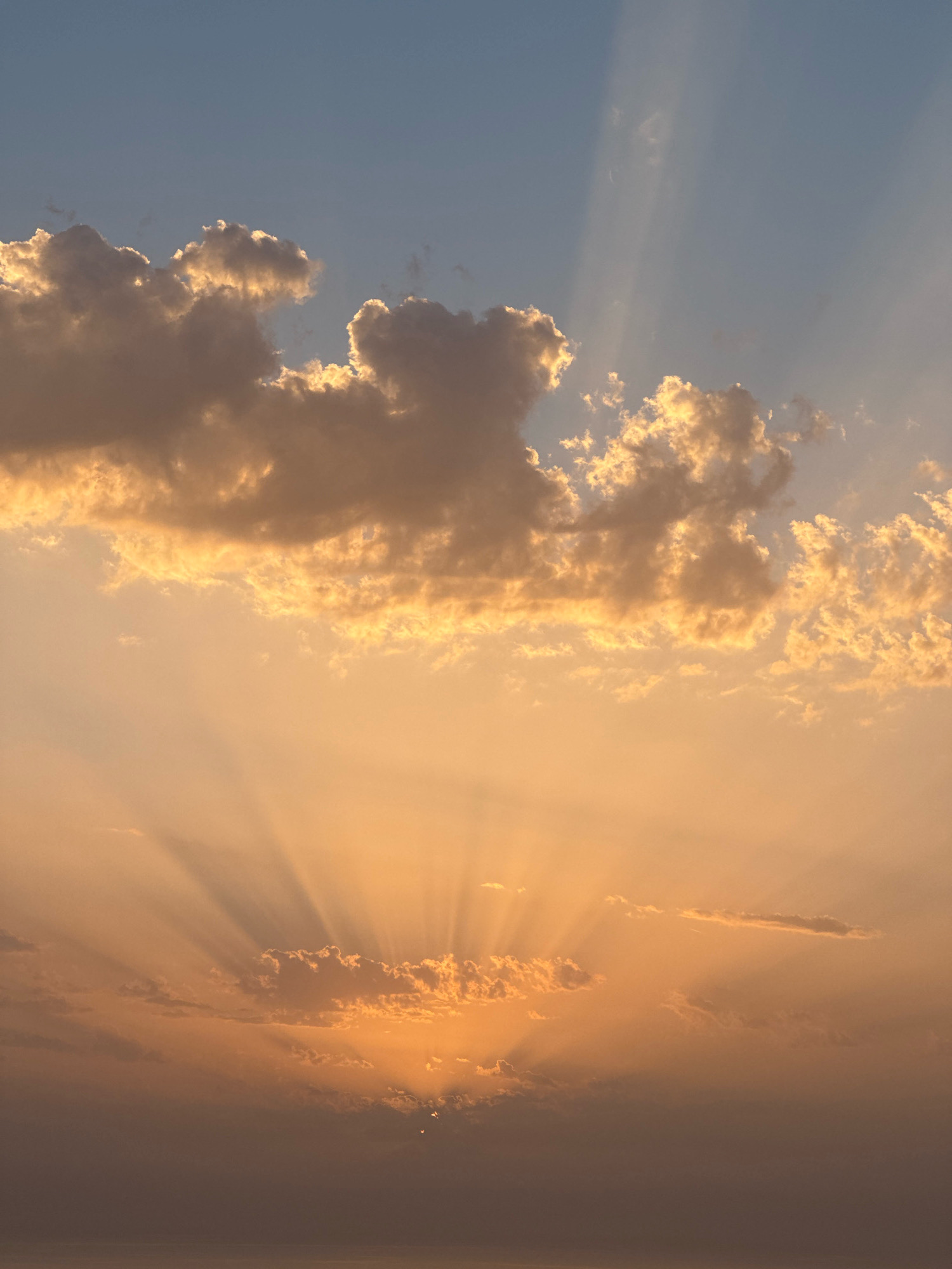 This is a vertical, eye-level, full shot of a sunset over the Adriatic Sea, viewed from Zagora, Montenegro. The sky is a gradient of colors, starting with a pale blue at the top and transitioning to warm oranges and browns closer to the horizon. Sunbeams pierce through scattered clouds, creating dramatic rays that fan out across the sky. The clouds themselves are illuminated by the setting sun, with some appearing bright white and others a dusky gray. The sea is visible at the bottom of the frame, a dark, calm expanse reflecting the sky's colors. There are no people or man-made objects visible in the image.
