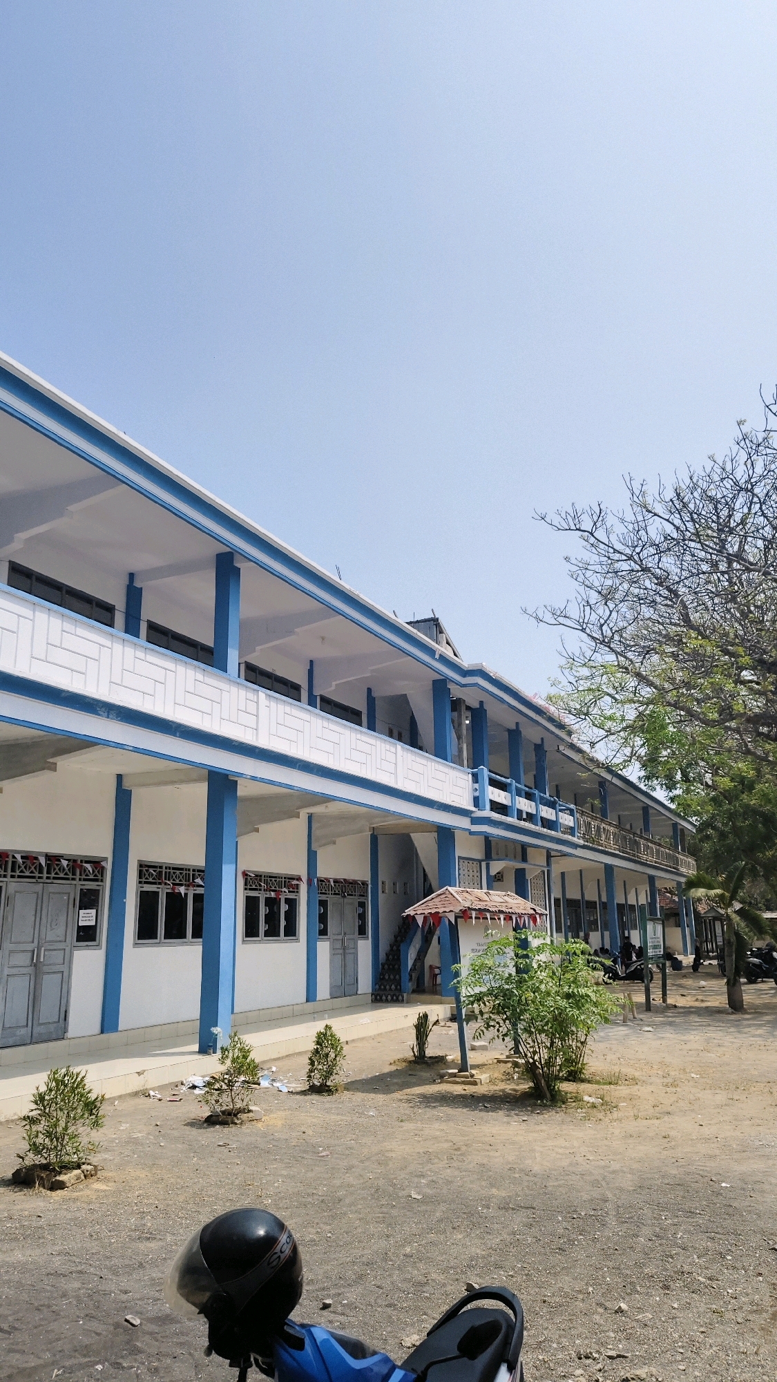 The image depicts the exterior of a two-story building, likely a school, in Pangpajung, Indonesia. The building is characterized by white walls with blue accents along the rooflines and structural columns. The second floor features a covered walkway or balcony with blue railings and decorative white latticework. Windows are visible on both floors, many with closed gray doors or shutters. Small green trees and shrubs are planted in front of the building, and the ground is covered with gravel and dirt. A blue motorcycle with a black helmet is parked in the foreground. In the background, a large tree with bare branches is visible, along with a clear blue sky. The scene suggests a bright, sunny day.