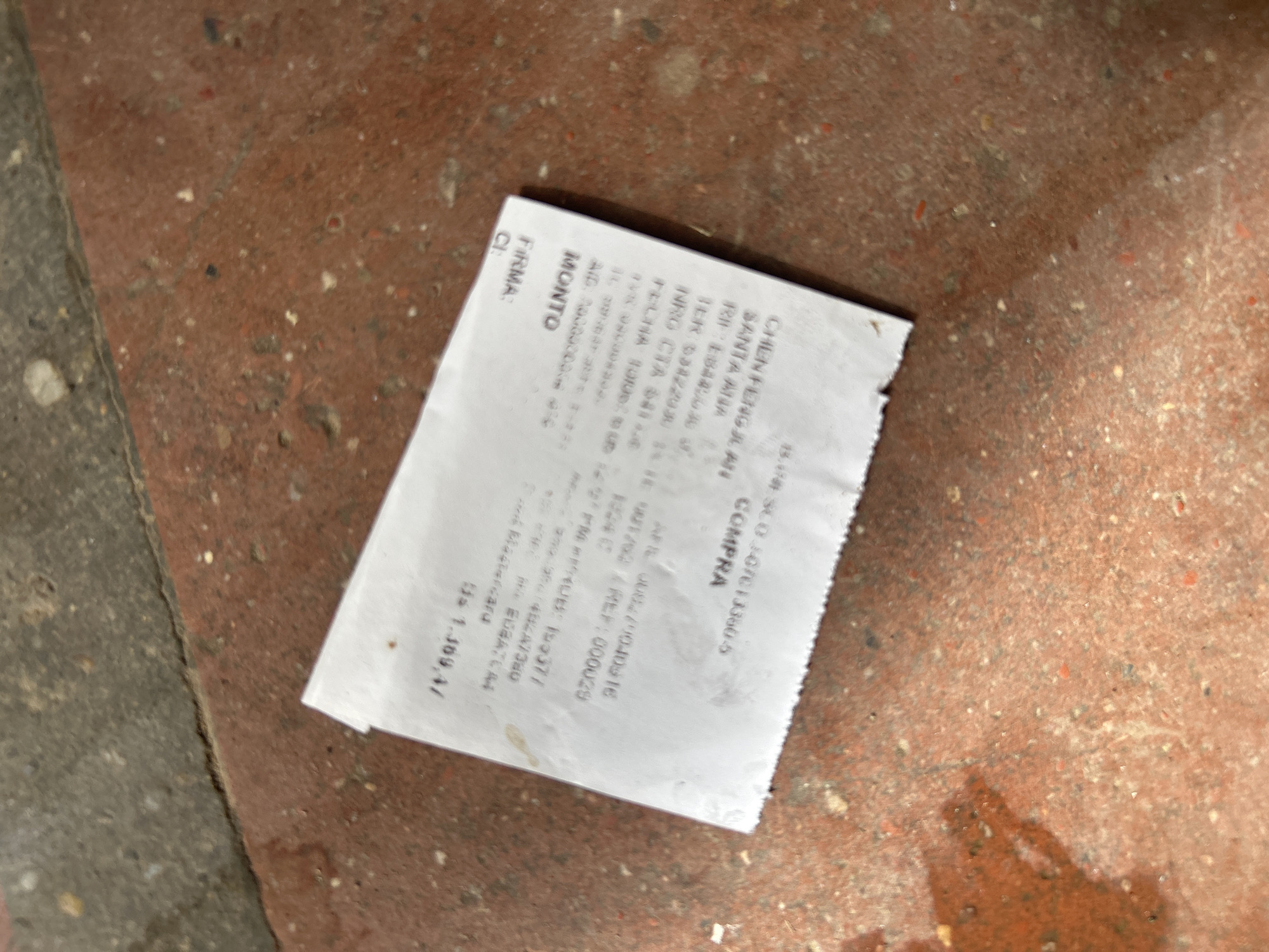 The image shows a crumpled receipt lying on a reddish-brown, textured surface, possibly concrete or asphalt with gravel. The receipt is white and has some dark text printed on it. The text is partially visible and appears to include financial information such as a total amount, a date, and possibly a store name. Some words like "MONTO," "FIRMA," and "COMPRA" are legible. The background is out of focus, but it appears to be an outdoor environment with a hint of shadow on the left side. The texture of the ground is rough with visible small stones and variations in color. The receipt itself is creased and slightly torn at the edges, suggesting it has been handled or exposed to the elements. The overall scene is low-angle, looking down at the receipt.