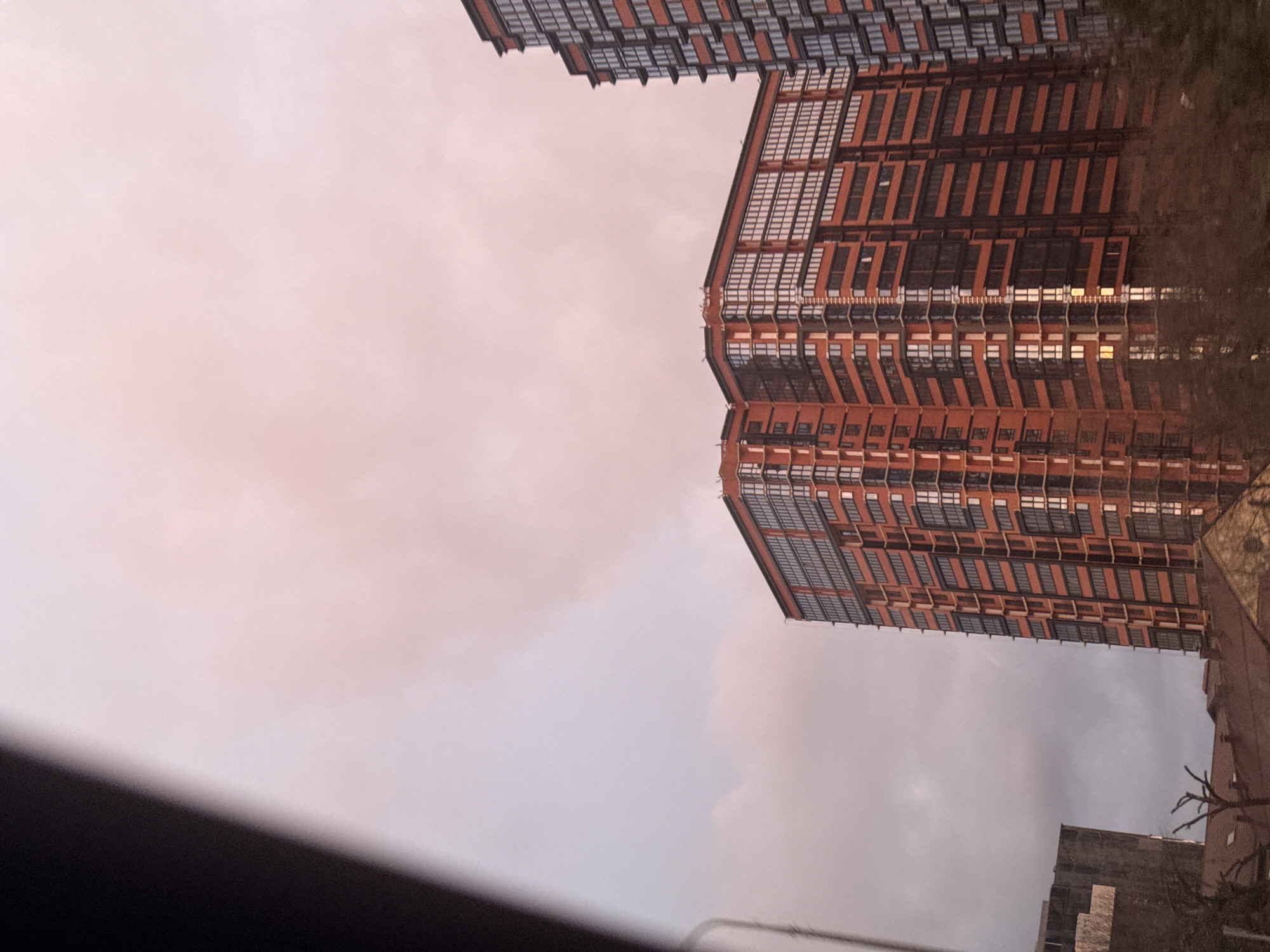 The image shows a low-angle view of tall buildings against a cloudy sky. The buildings appear to be residential or office towers with a grid of windows and a reddish-brown facade. The sky is overcast with varying shades of gray and hints of pink. The photo seems to have been taken from a lower level, possibly a street.