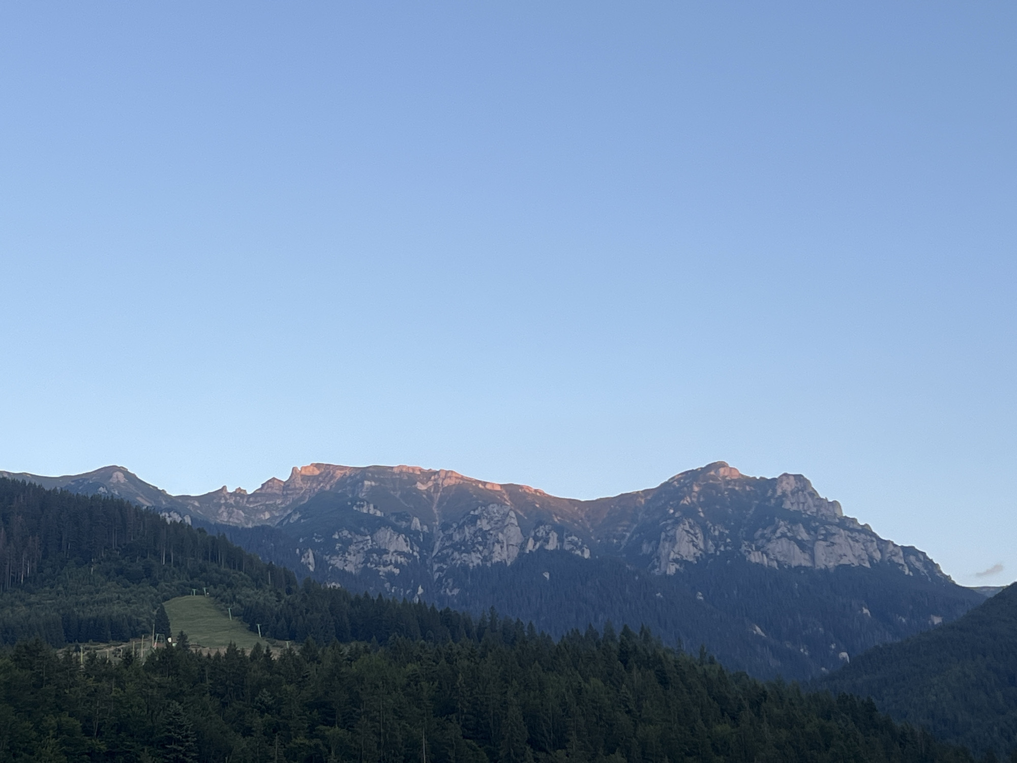 This is a wide landscape photograph of a mountain range under a clear blue sky. The mountains are covered in dark green conifer forests that rise to meet rocky, jagged peaks. The setting sun casts a warm, golden light on the highest points of the mountain range, creating a stark contrast with the shadowed slopes below. A ski slope with a few ski lifts is visible on the green hillside in the mid-ground, suggesting the location is a mountain resort. The foreground is dominated by dense trees. The overall scene depicts a serene and majestic natural landscape.