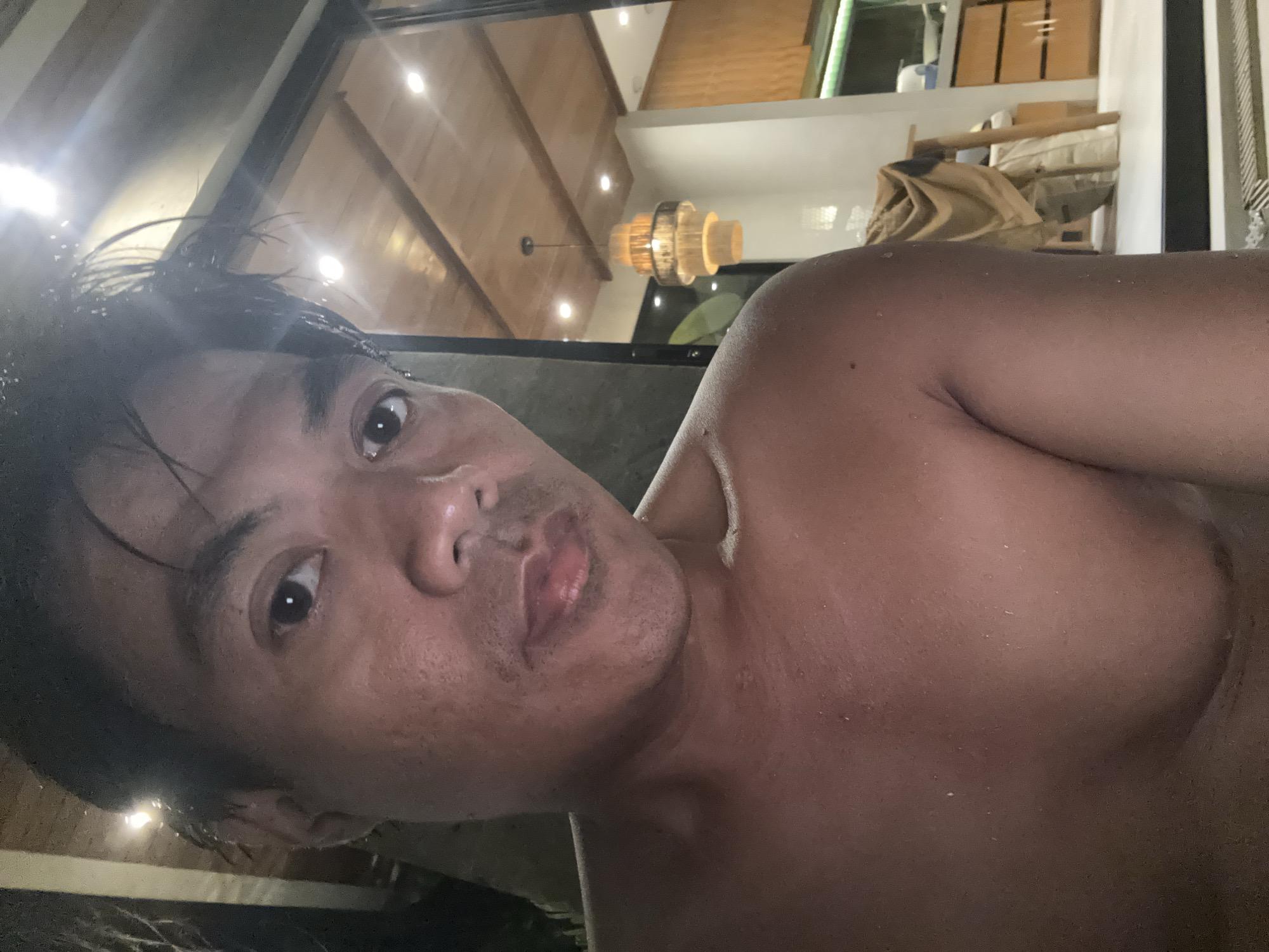 The image shows a shirtless individual with wet hair in an interior setting. The person has dark hair and is looking directly at the camera. The background includes a wooden ceiling with recessed lighting and a chandelier. A partially visible interior space with furniture is seen behind the person. The picture appears to have been taken indoors, possibly in a residential location or a resort-style setting. The location appears to be Tumbak Bayuh, Indonesia.