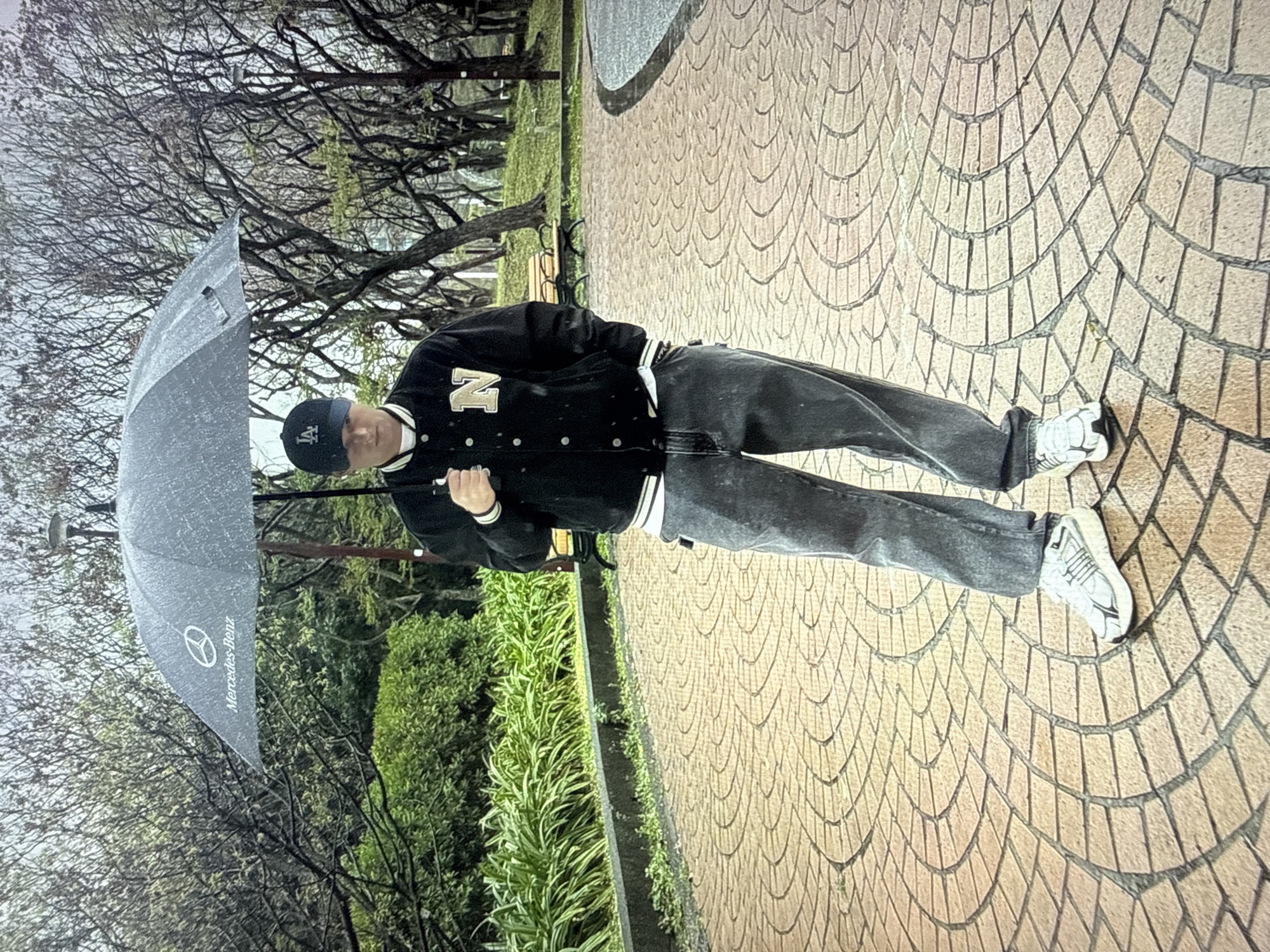 A person stands in a park on a rainy day holding a Mercedes-Benz umbrella. The person is wearing a black jacket with a white "N" on the chest, a white shirt, dark gray jeans, and white sneakers. The ground is paved with curved patterns of light brown bricks. In the background, there are lush green bushes, bare trees, and a park bench. The overall atmosphere is damp and cool, consistent with a wet day in a city park.
