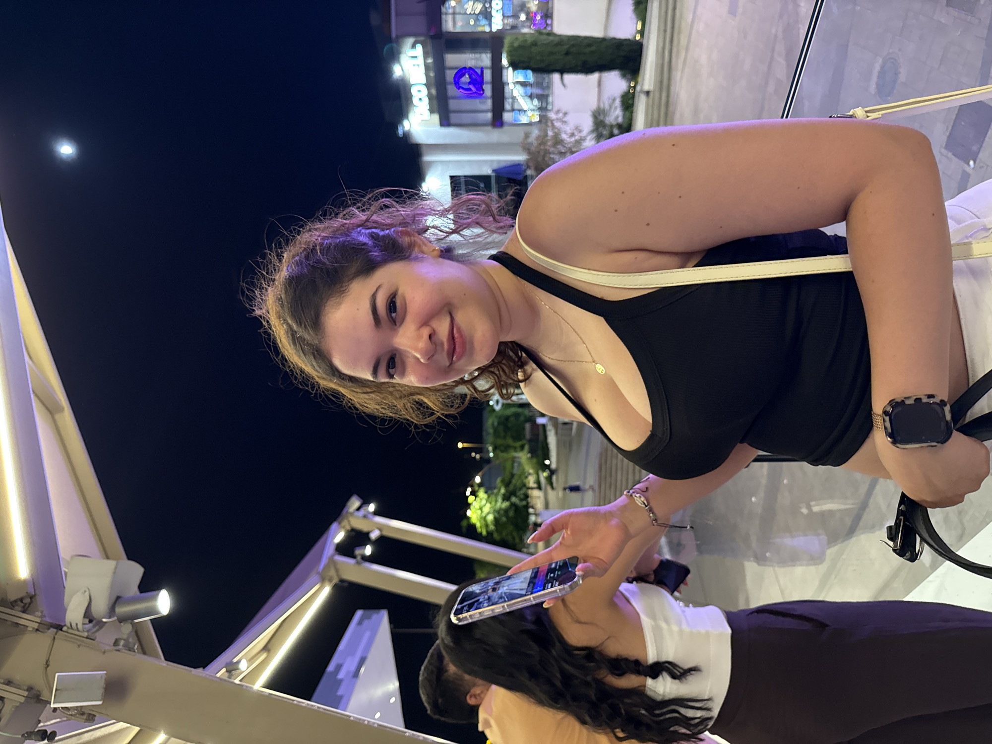 A young woman with curly brown hair and light skin is posing for a picture at night. She is wearing a black tank top, a gold necklace, and a bracelet. She is holding a smartphone in her left hand. In the background, there is a Taco Bell restaurant, trees, and streetlights. The moon is visible in the dark sky. The woman is standing on a balcony or walkway with a glass railing. Two other people are partially visible in the background, one wearing a white top and dark pants, and another wearing an orange shirt. The scene appears to be an outdoor dining or entertainment area.