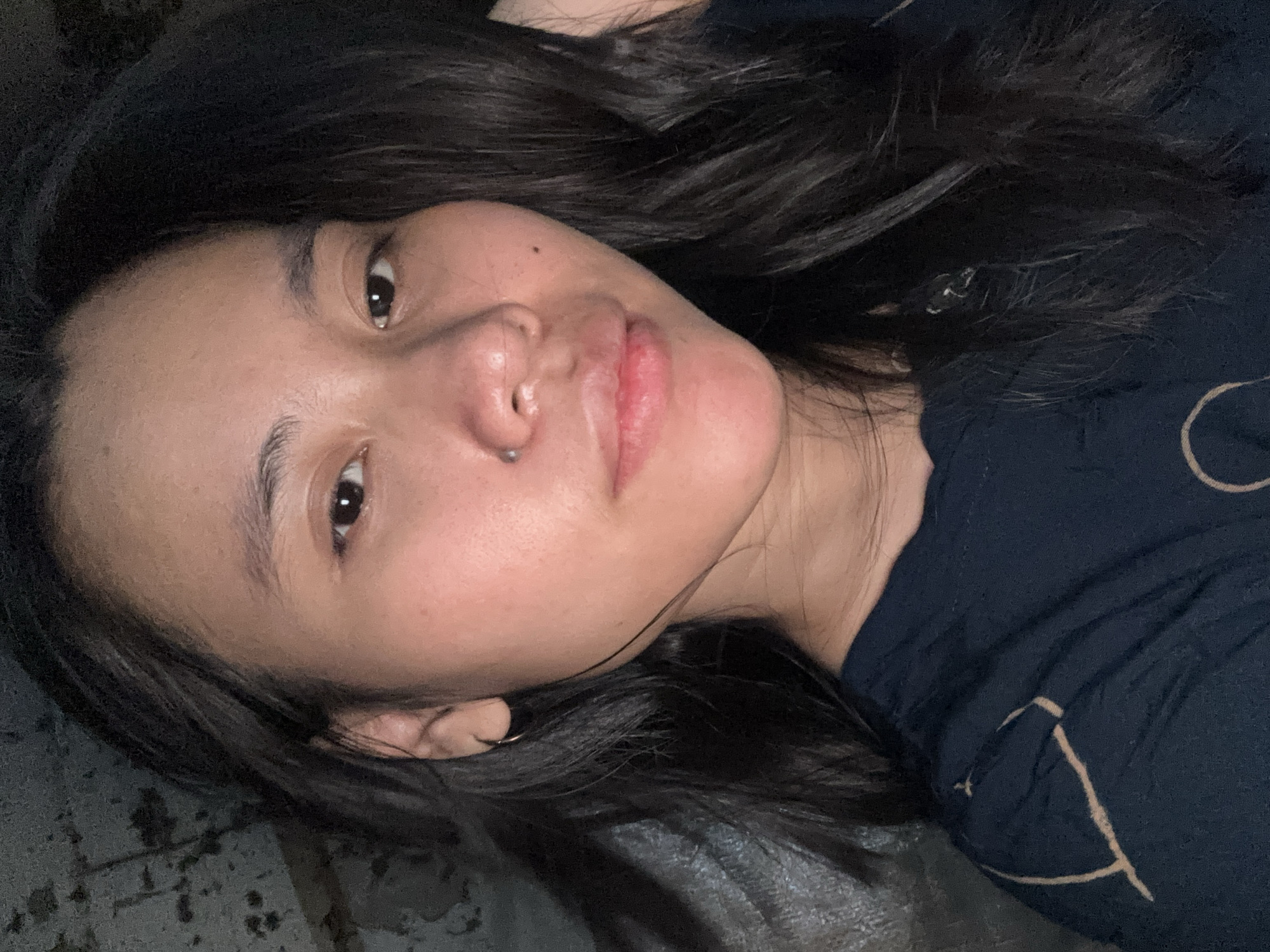 This media file features a close-up, rotated shot of a young person lying down. Their face is the primary subject, with dark hair framing their head and falling across their shoulders and shirt. The person has light brown eyes, dark eyebrows, and a nose piercing. Their lips are slightly parted, and their skin appears to have some subtle blemishes. The overall lighting suggests an indoor setting, possibly at night or in a dimly lit room, as there are no strong indicators of natural light. The person is wearing a dark blue t-shirt with a light brown, abstract pattern. The background is dark and textured, appearing to be a surface like a bed or couch. The image conveys a relaxed or casual mood, with the person looking towards the camera. There is no visible text in the image. The location context provided is East Jakarta, Indonesia.