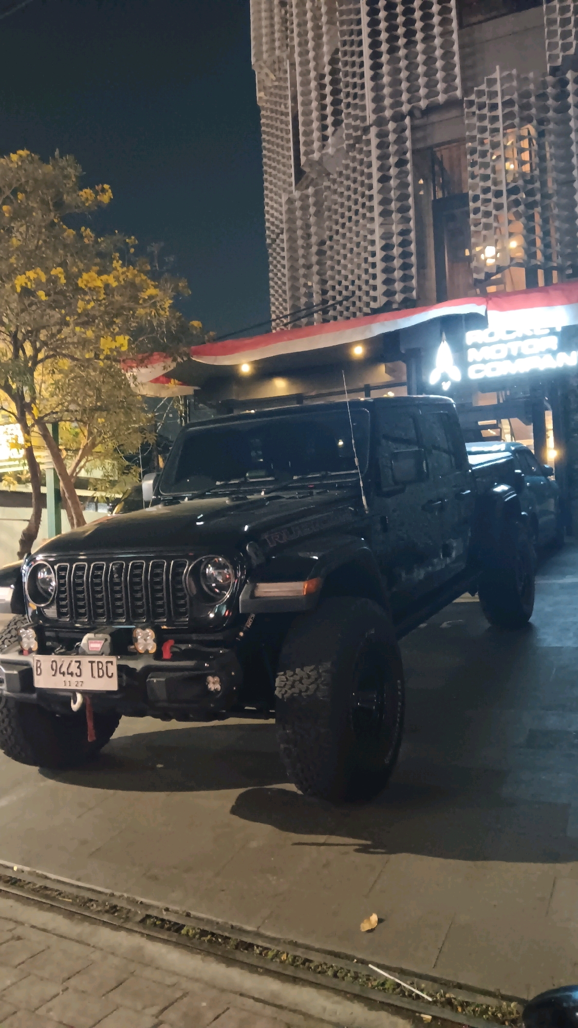 A black Jeep Wrangler Rubicon is parked on a sidewalk in front of a building at night. The building has a unique facade with a patterned screen. A tree with yellow flowers is visible to the left of the Jeep. The license plate on the Jeep reads "B 9443 TBC". A sign on the building is partially visible and appears to say "ROCKET MOTOR COMPANY". The scene is consistent with an urban environment in South Jakarta, Indonesia.