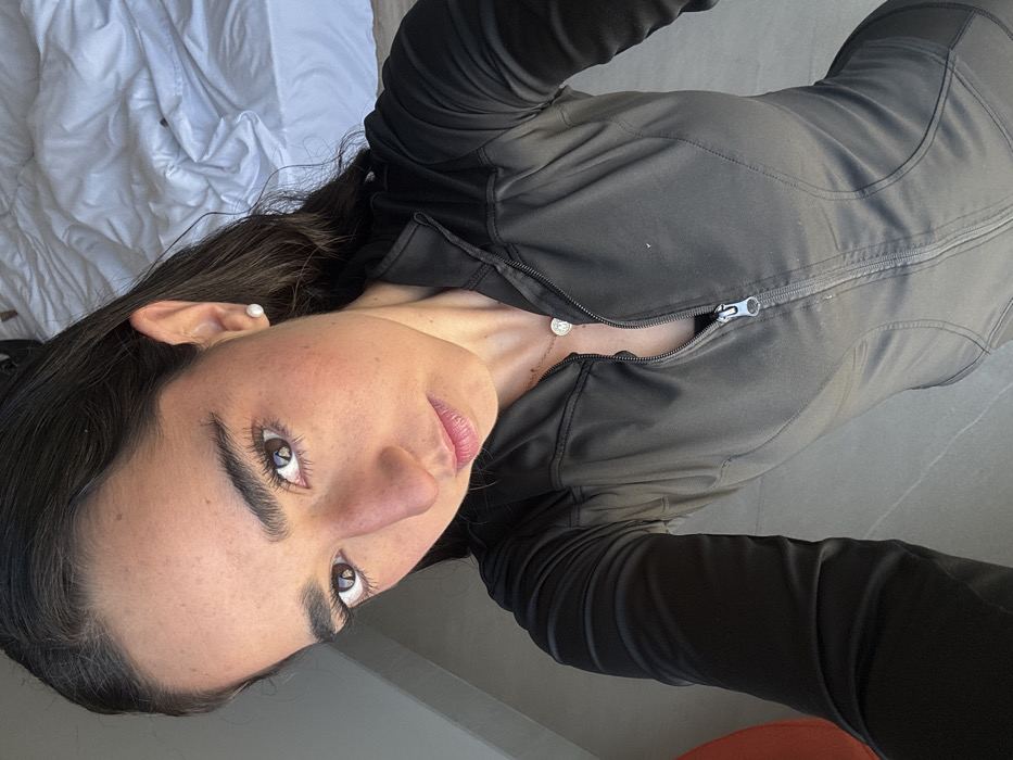 A female subject is depicted in a high-angle, close-up indoor portrait. She has dark hair, brown eyes, and is looking directly at the camera. She wears small, round white pearl stud earrings and a silver-toned necklace with a circular medallion pendant. Her attire consists of a dark gray or black long-sleeved athletic top or jacket with contrasting black fabric sections, featuring a partially unzipped front zipper. The background includes a rumpled white bed to the right, a light-colored floor, and a vertical gray structure to the left. A blurred orange object is present in the lower left corner. The scene location is Unknown, Unknown.