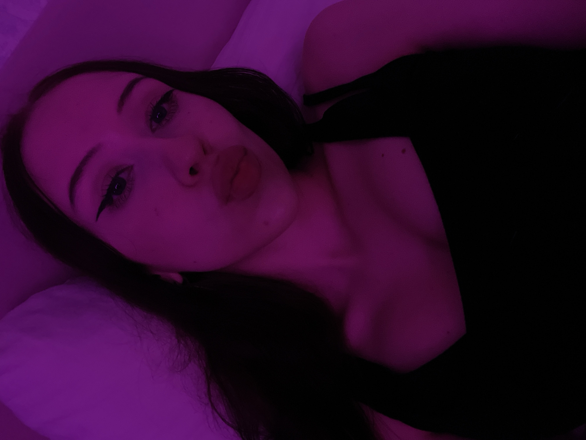 The image shows a close-up, chest-up view of a young woman lying down, illuminated by purple light. She has long, dark hair that falls around her shoulders and a black tank top. Her eyes are open and looking at the camera, with dramatic winged eyeliner. Her lips are slightly parted, in a pout. The background is a soft, blurred purple surface, possibly a pillow or bedding. There is no visible indication of the city of Votkinsk, Russia, or any urban environment. The scene is indoor and private.