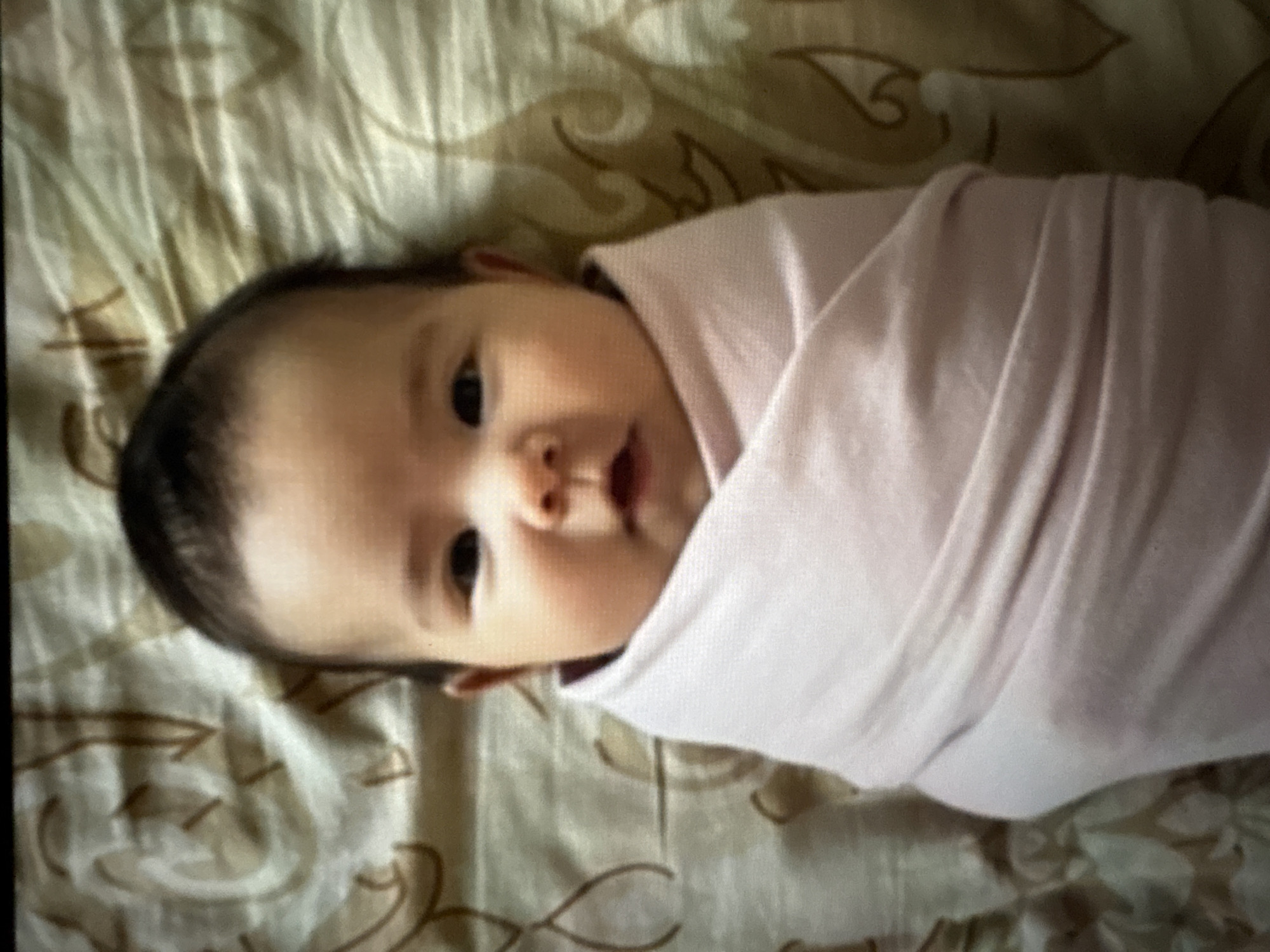 A baby is swaddled in a light pink blanket on a bed with a beige patterned sheet. The baby is looking directly at the camera.
I do not have information about the city.