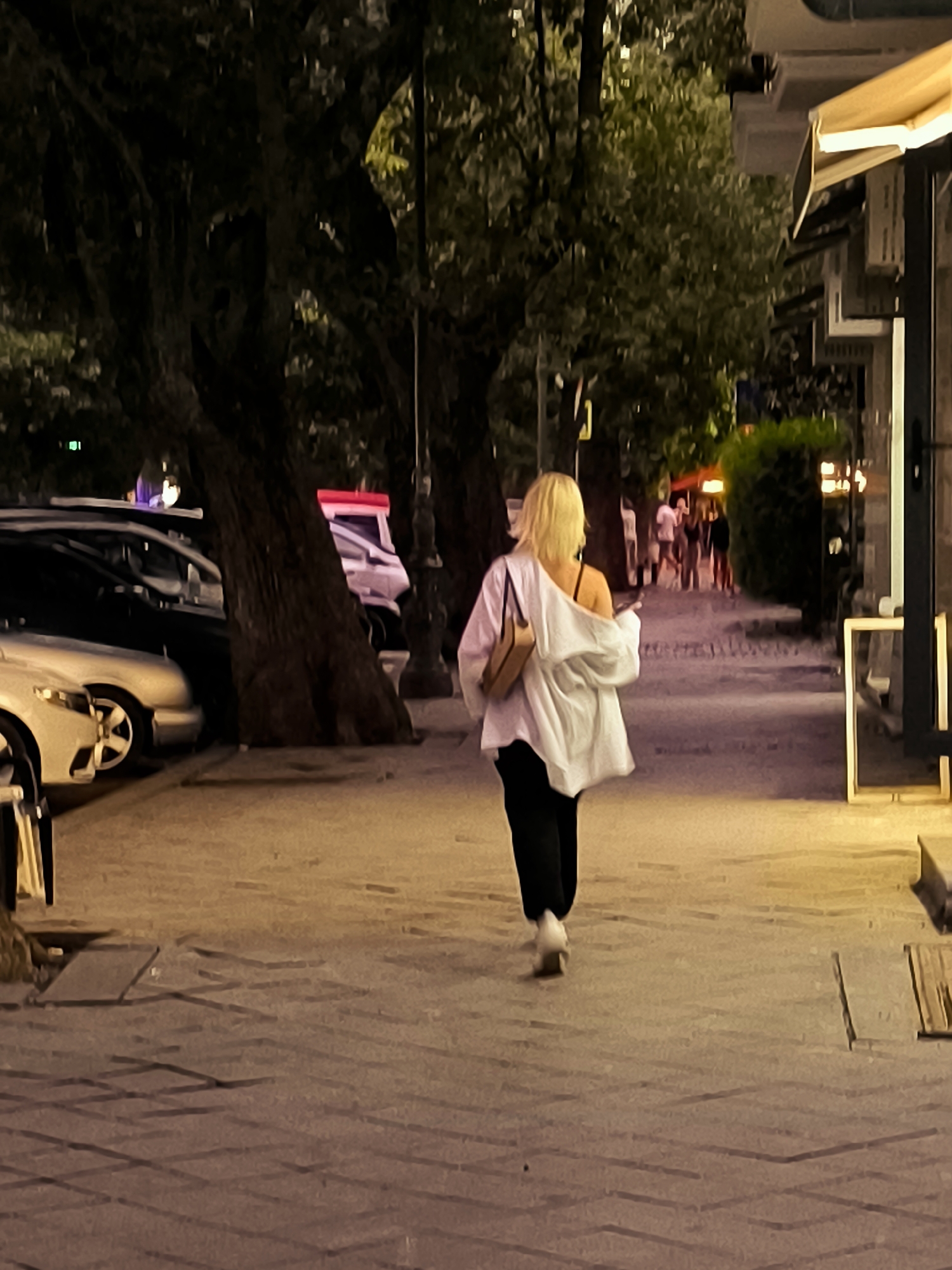 A woman with blonde hair and a white shirt is walking away from the camera on a paved sidewalk at night. She is carrying a tan purse over her shoulder and is wearing black pants. In the background, several cars are parked on the left side of the street, and a few people are visible walking in the distance. The scene appears to be a street in Batumi, Georgia, likely near a commercial area given the presence of shops and awnings. The lighting suggests it is evening or nighttime.