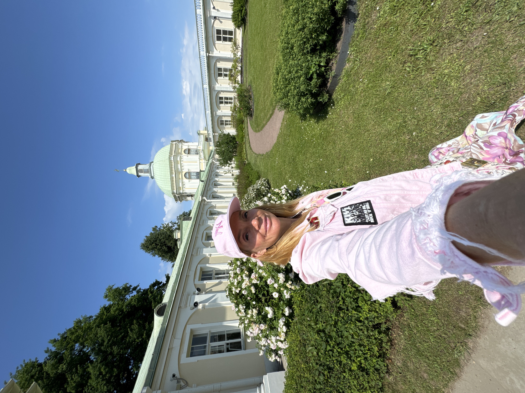 A woman with blonde hair, wearing a pink baseball cap and a distressed pink denim jacket, smiles for a selfie outdoors. She is positioned in the foreground with a grand, light-colored building and lush green landscaping behind her. The building features a prominent green dome and a long facade with numerous windows and arched entrances. A winding path cuts through a manicured lawn, bordered by dense green bushes and flowering plants. The sky is a clear, bright blue, suggesting a sunny day. The overall atmosphere is bright and cheerful, with the woman appearing happy and relaxed. The building's architectural style hints at a historical or governmental site, consistent with the Lomonosov, Russia context. Visible text on the woman's jacket includes a patch that reads "ROCK & ROLL" and another with figures that appear to be musicians. The time of day is likely midday, given the strong sunlight and shadows.