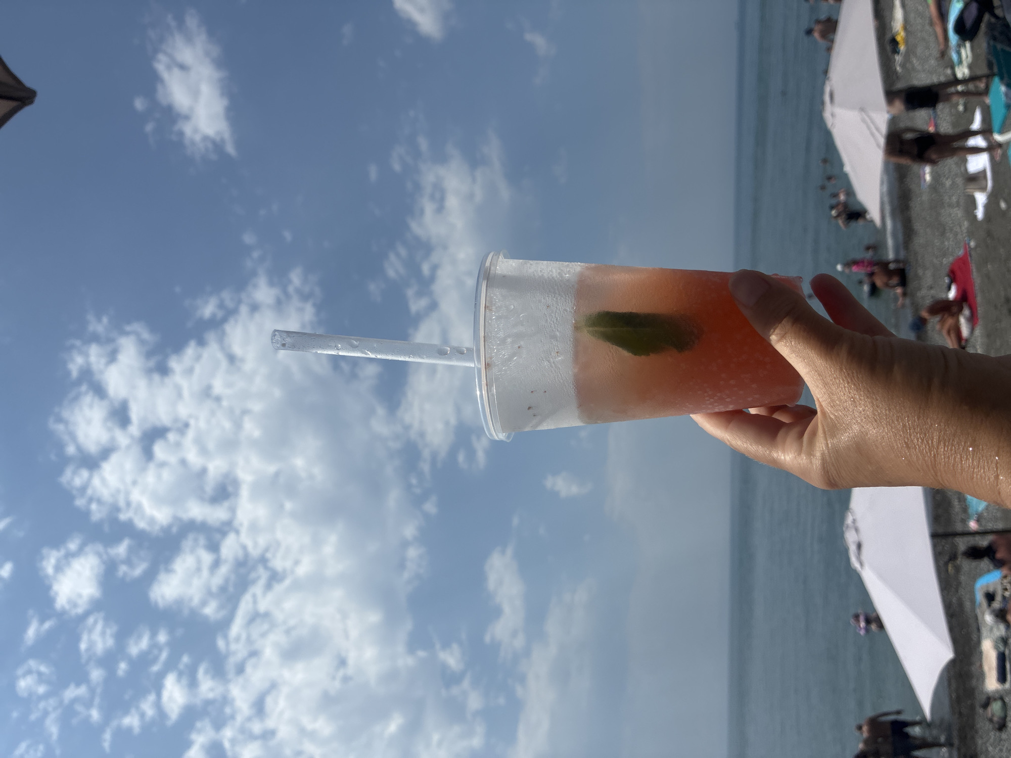 An overcast sky with scattered white clouds is visible above a beach scene. In the foreground, a person's hand holds a clear plastic cup filled with a pink beverage, a clear straw, and a green leaf. The cup is frosted, suggesting it contains a cold drink. The beach is covered in pebbles, and several white umbrellas are open. People are visible in the background, some standing in the shallow water of the ocean, others relaxing on the beach. The location is identified as the city of Sirius, Russia.