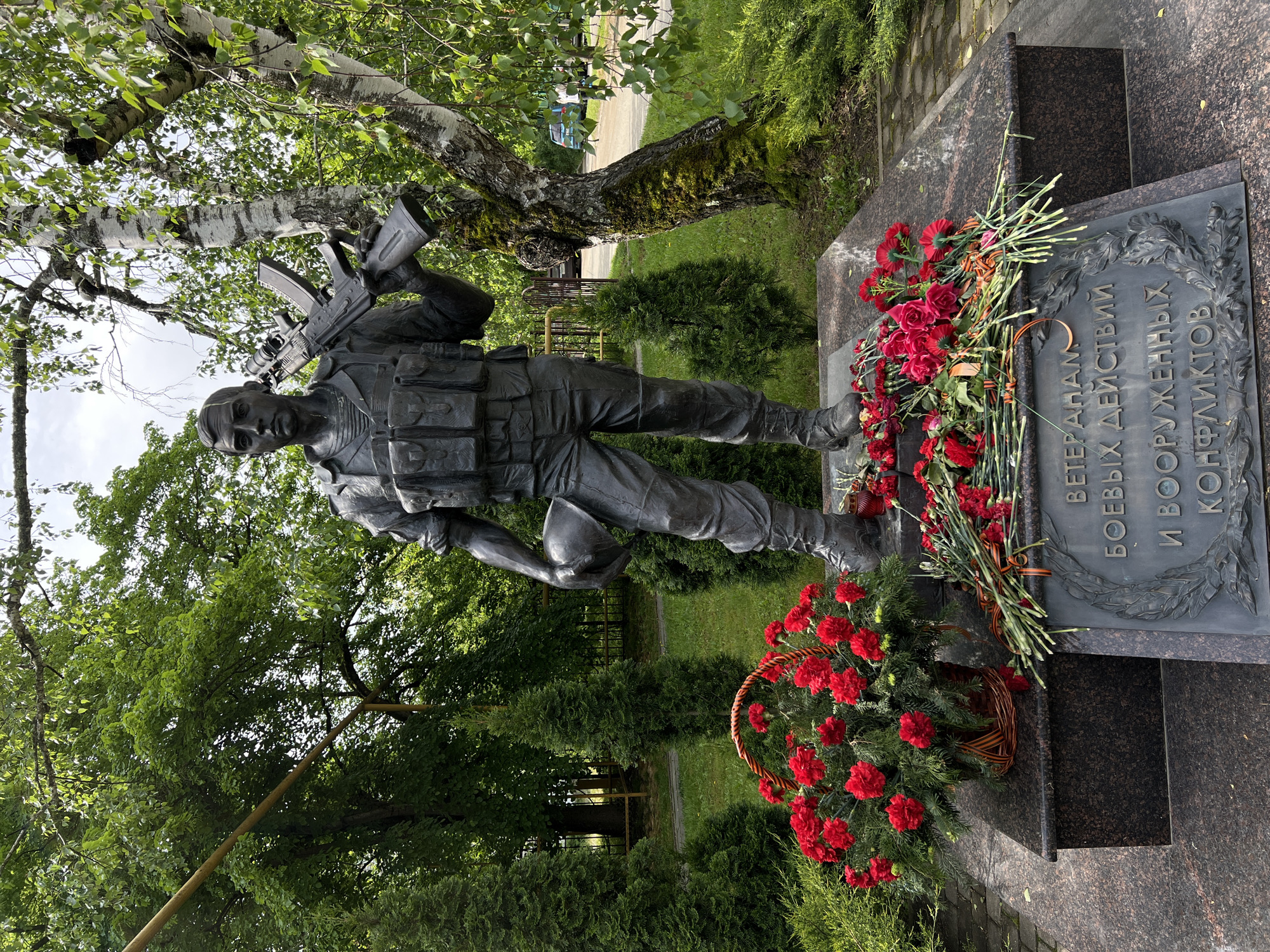 The image shows a bronze statue of a soldier holding a rifle and helmet, placed on a granite memorial with Cyrillic inscription. Red flowers, some in a basket and others laid as bouquets with orange and black ribbon, adorn the base of the memorial. The statue and memorial are located outdoors, surrounded by trees and shrubs. The location is Goryachiy Klyuch, Russia.
