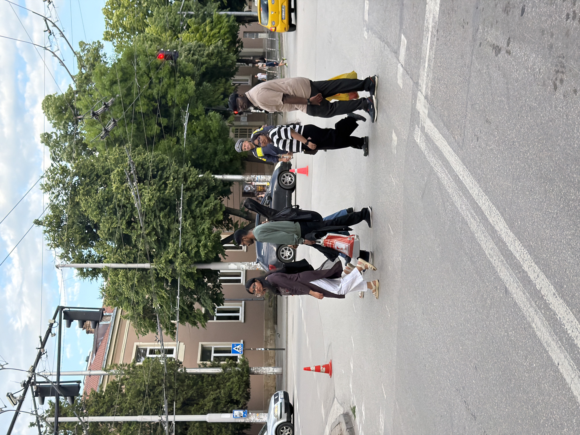 The image depicts a street scene in Pleven, Bulgaria, featuring a group of four people crossing a street. One man wears a police uniform and appears to be assisting the other three across the street. The first man is wearing a tan shirt and black pants, the second man is wearing a black and white striped shirt with black pants, the third man is wearing a light green shirt and jeans, and the woman in the group is wearing a maroon shirt and white pants. They are crossing at an intersection with a traffic light that is red. The street is marked with white lines indicating lanes. Several cars are visible, including a yellow taxi in the background, and one parked along the sidewalk. A building with multiple windows can be seen on the left side of the image. The background consists of trees, utility poles, and buildings.