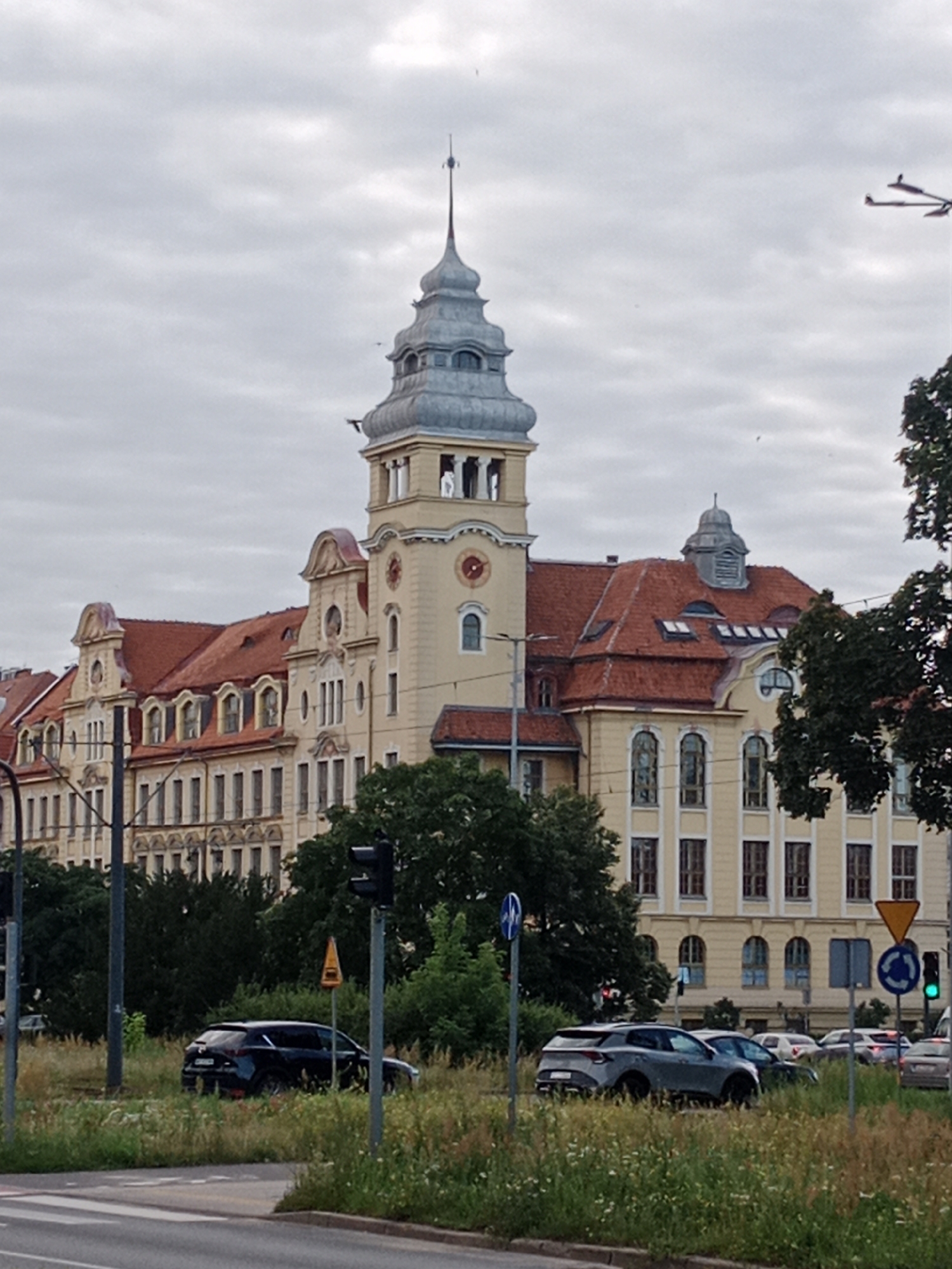 The image captures a street view in Bydgoszcz, Poland, featuring a large, historic building with a prominent tower and a red-tiled roof. The building is light yellow with multiple windows and ornate architectural details. In the foreground, there are cars parked or driving on the street, and a grassy median with wildflowers separates the road from the building. Traffic lights and street signs are also visible. The sky is overcast. The specific building is the Polish Post building (Poczta Polska), located at Gdańska Street 15 in Bydgoszcz.