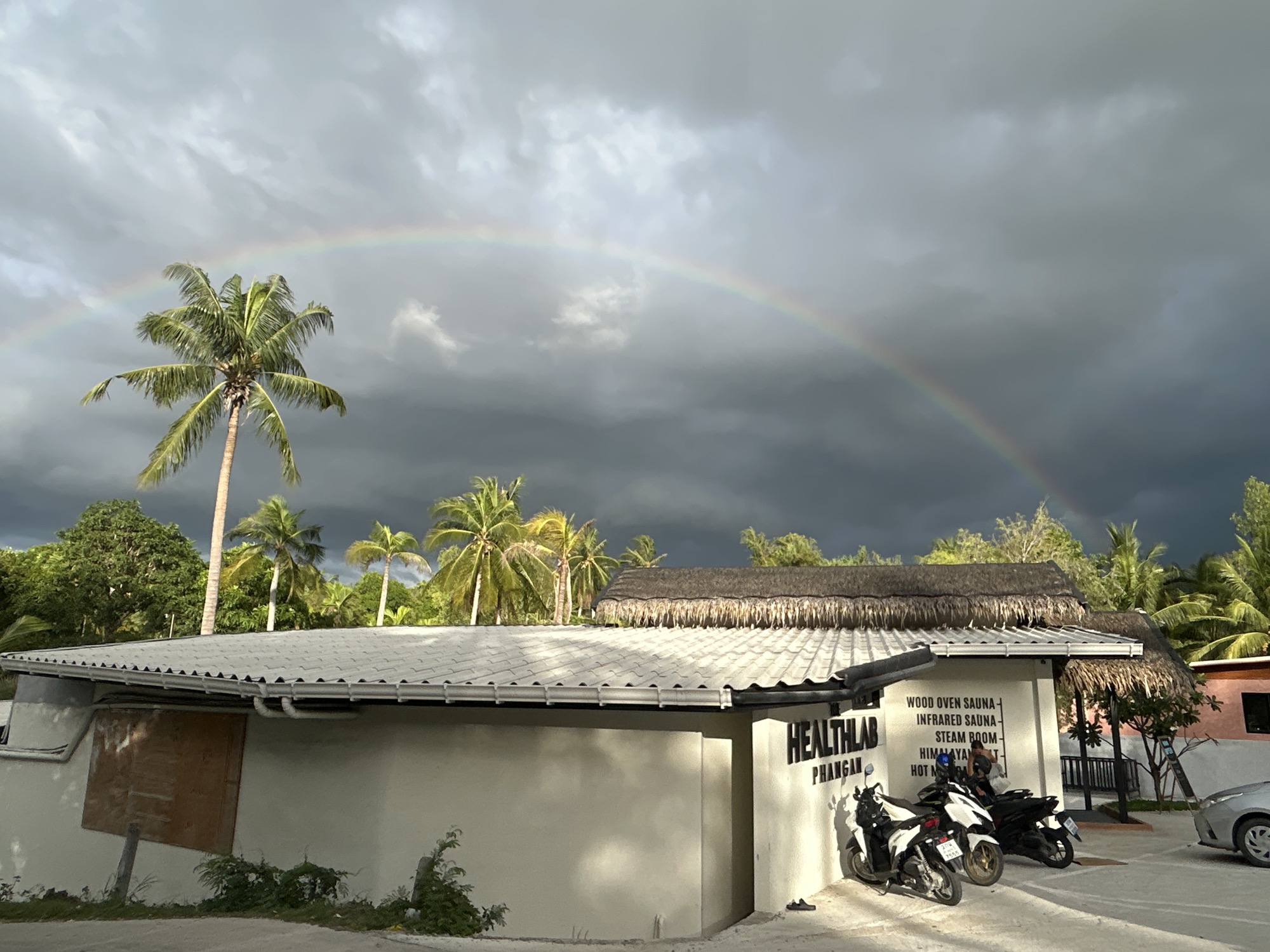 The image depicts a scene in Baan Hin Kong, Thailand, under a cloudy sky with a prominent rainbow. The architecture features a white building with a gray tiled roof and a thatched-roof extension. The sign on the building reads "HEALTHLAB PHANGAN", followed by "WOOD OVEN SAUNA, INFRARED SAUNA, STEAM ROOM, HIMALAYAN...", although the rest is not visible. In front of the building, three motorbikes are parked. A car is partially visible on the right side of the image. The background includes various palm trees and lush vegetation, typical of a tropical setting. The overall environment suggests a combination of natural and commercial elements.