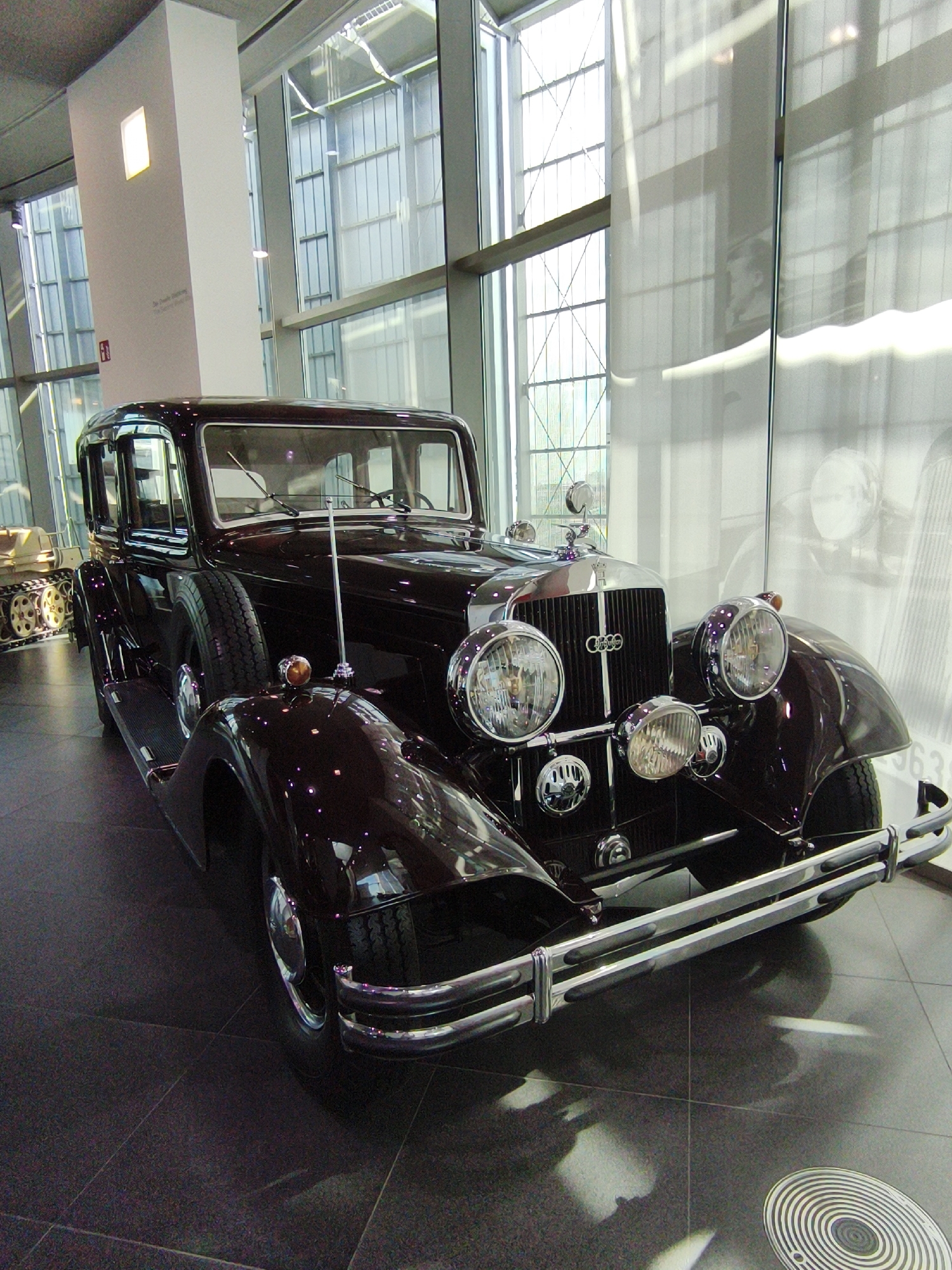 An antique dark red Audi sedan is displayed in a museum setting, likely the Audi Forum in Ingolstadt, Germany, given the architectural style of the building with its large glass windows and industrial interior. The car is positioned centrally in the frame, viewed from a low angle, highlighting its classic design. The vehicle features prominent round headlights, a chrome grille with the Audi rings emblem, and a spare tire mounted on the side. To the left of the sedan, a portion of a tracked vehicle, possibly a historical military vehicle, is visible. The floor is a dark, polished stone, reflecting the ambient light and the car. The background consists of large windows with a criss-cross structural pattern, suggesting a modern building. There are no people in the image.