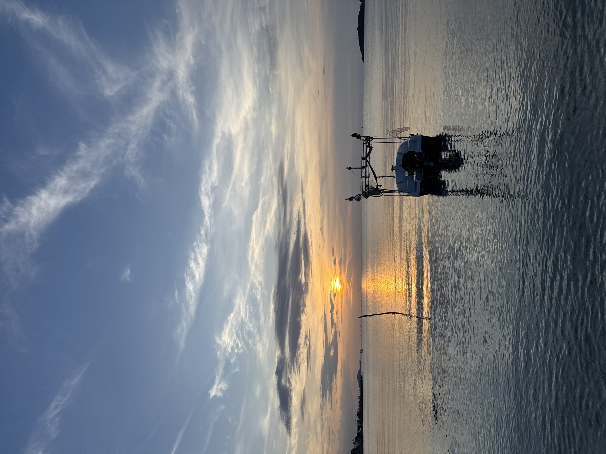 A boat is anchored in the calm waters of Ko Samui District, Thailand, at sunset. The sun, a bright orange orb, is low on the horizon, casting a golden glow on the water and reflecting off the sky. Wispy clouds drift across the blue expanse, tinged with pink and orange hues from the setting sun. The water is a deep blue, with gentle ripples creating a textured surface. In the foreground, a single wooden post juts out of the water, its reflection shimmering on the surface. The boat, a small white vessel with a dark outboard motor, is positioned in the mid-ground. It has a metal frame structure on its deck, possibly for fishing equipment, and two nets are visible hanging from the sides. In the distance, a faint silhouette of land and buildings can be seen on the left, and a small island or landmass is visible on the right horizon. The overall scene is tranquil and serene, capturing the peaceful beauty of a sunset over the ocean.