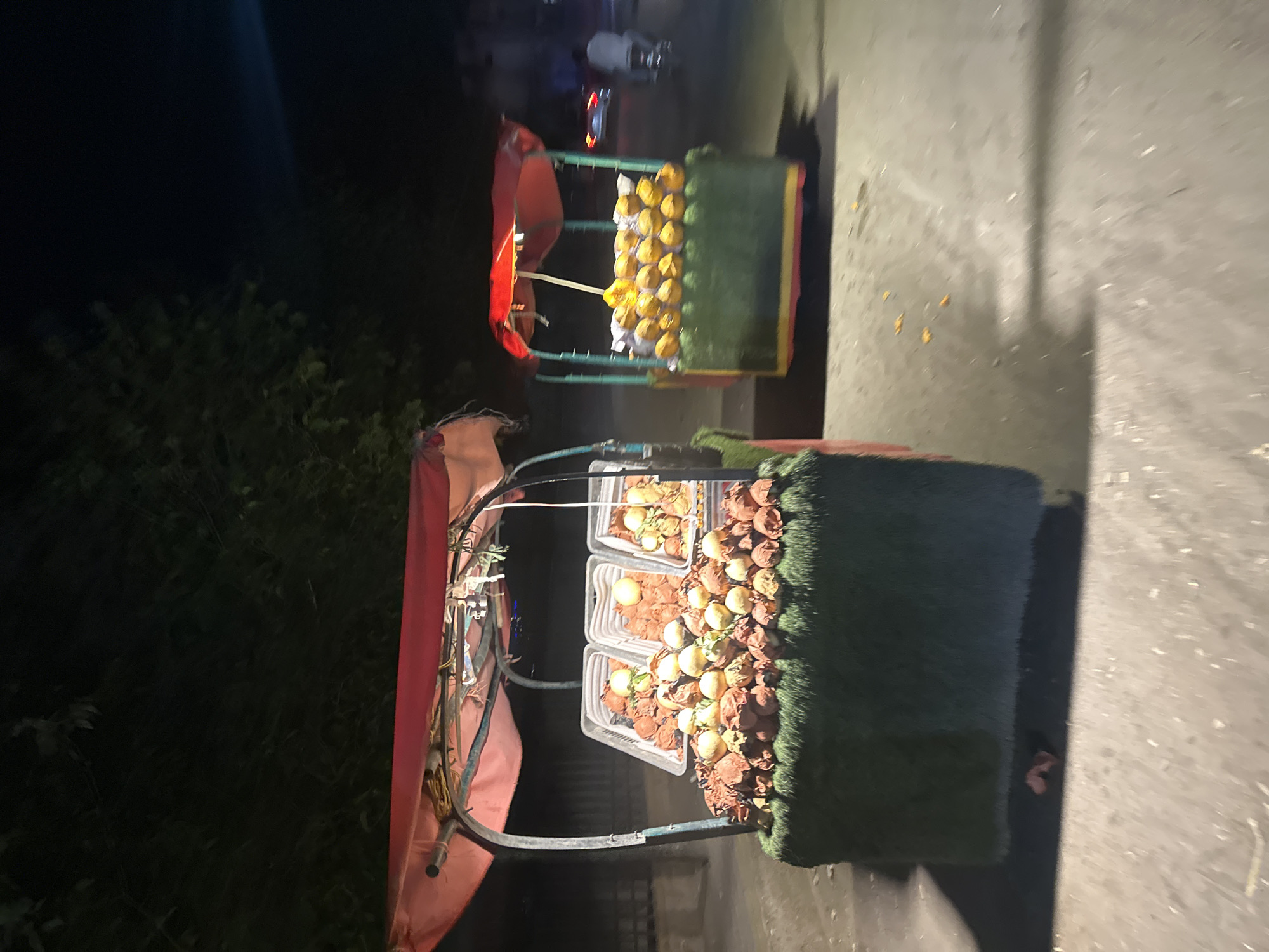 Captured at night in Quetta City Tehsil, Pakistan, the image depicts two street vendor carts. The closest cart has three plastic containers filled with fruit, surrounded by dried leaves and covered with artificial grass. The cart also has a red canvas top. The second cart in the background displays yellow fruits arranged in rows, also underneath a red canvas. A motorcycle passes in the background, and the ground is paved. Vegetation is visible on the left of the frame.