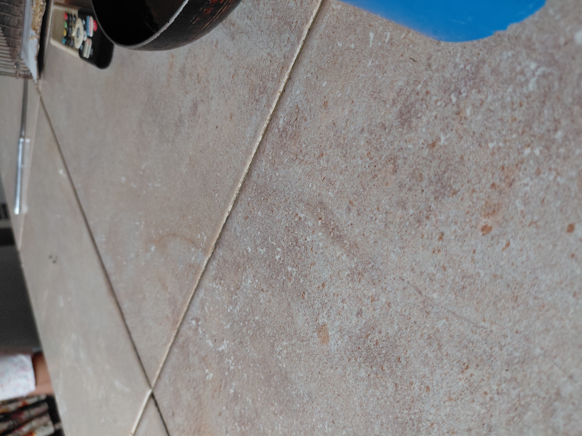 The image presents a close-up view of a light-colored tiled surface, likely a floor or countertop, within an indoor setting in Santa Ana, Venezuela. The tiles are a muted beige-brown with a mottled, textured appearance, featuring scattered white and darker brown speckles. Visible grout lines divide the tiles, and a fine, white, powdery residue, possibly dust or dried spillage, is distributed across the surface.
In the upper left portion of the frame, household items are partially visible. A black remote control with several indistinct colorful buttons rests near a dark, circular object that resembles the rim or bottom of a pan or pot. Adjacent to these, a slim, reflective object, possibly made of metal or glass, protrudes into view. Toward the upper right, a vibrant blue object or surface curves into the shot, its specific form or purpose unclear. No particular activity is depicted, but the items suggest a domestic environment. There is no decipherable text visible on any of the objects. The time of day and weather cannot be determined from the image.
