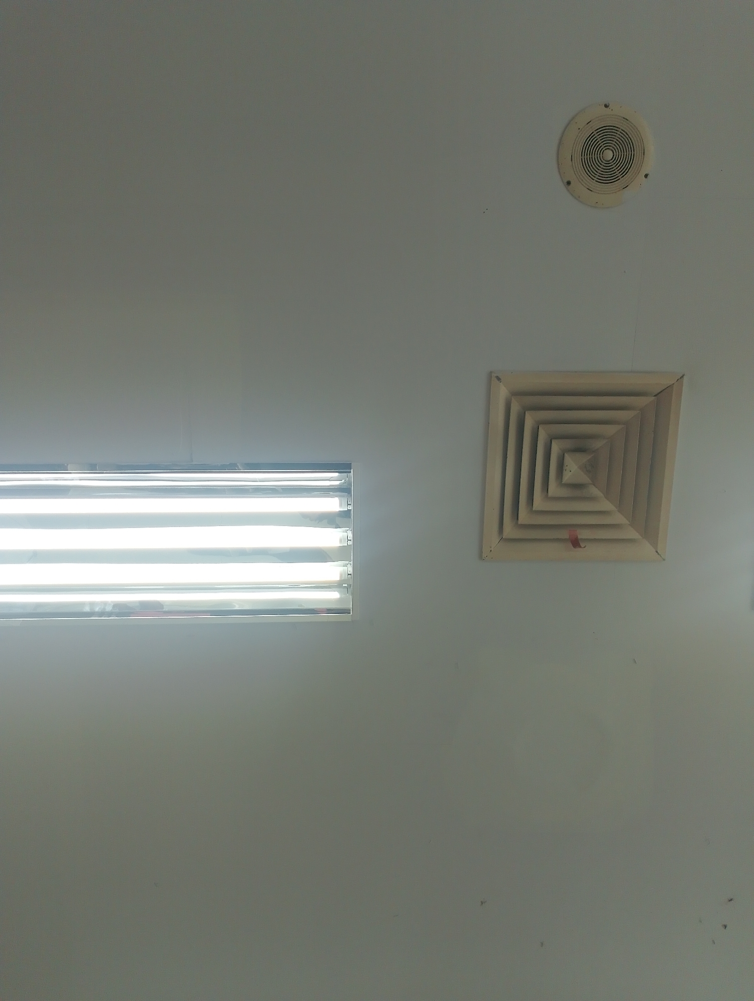 An interior shot of a room with a light gray ceiling is captured. The ceiling features two air vents and a long, rectangular fluorescent light fixture. One air vent is circular and located in the upper right quadrant of the image. It is cream-colored with a concentric ring design. The second air vent is square and has a stepped, recessed design, also cream-colored. This vent is situated to the right of the center of the image. A thin, red ribbon is attached to the bottom of the square vent. The light fixture, a long rectangle with multiple fluorescent tubes, spans the left side of the image, emitting a bright, white light. The overall scene suggests a modern, utilitarian space, possibly an office or a commercial establishment. The image does not provide specific details to determine its exact location within Batam, Indonesia, but the lighting and air vents are common features found in such environments.