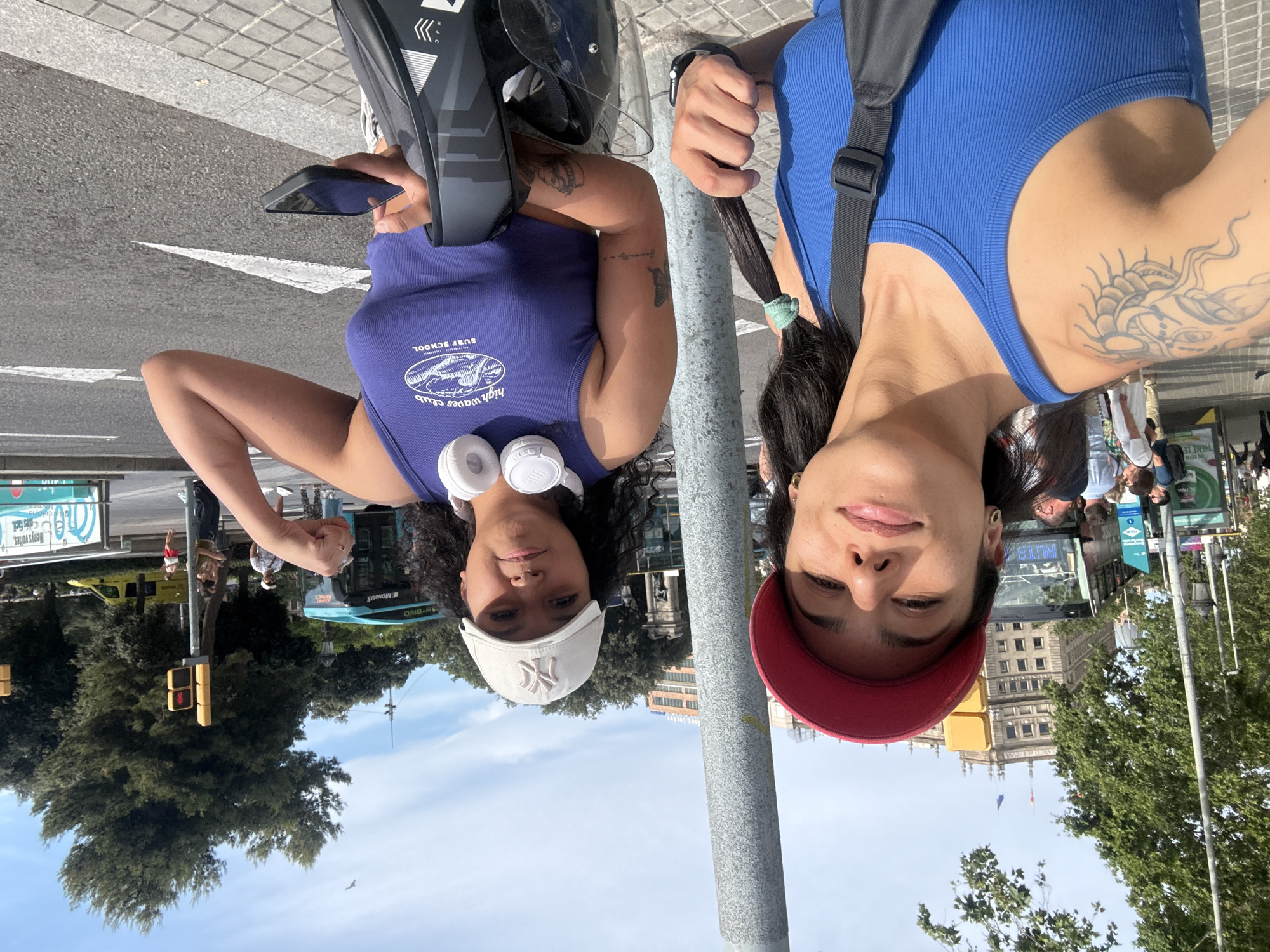Two women are standing on a sidewalk next to a busy street with traffic and pedestrians. The woman on the left is wearing a red baseball cap and a blue tank top. She has a tattoo on her left arm and is holding her braided hair with a green scrunchie. The woman on the right is wearing a white baseball cap and a purple tank top with a graphic that says "high waves club surf school". She has headphones around her neck and is flexing her right arm. She is holding a smartphone in her right hand. In the background, there are buses, cars, buildings, and trees. The sky is blue with some clouds. The scene appears to be a sunny day in an urban environment.