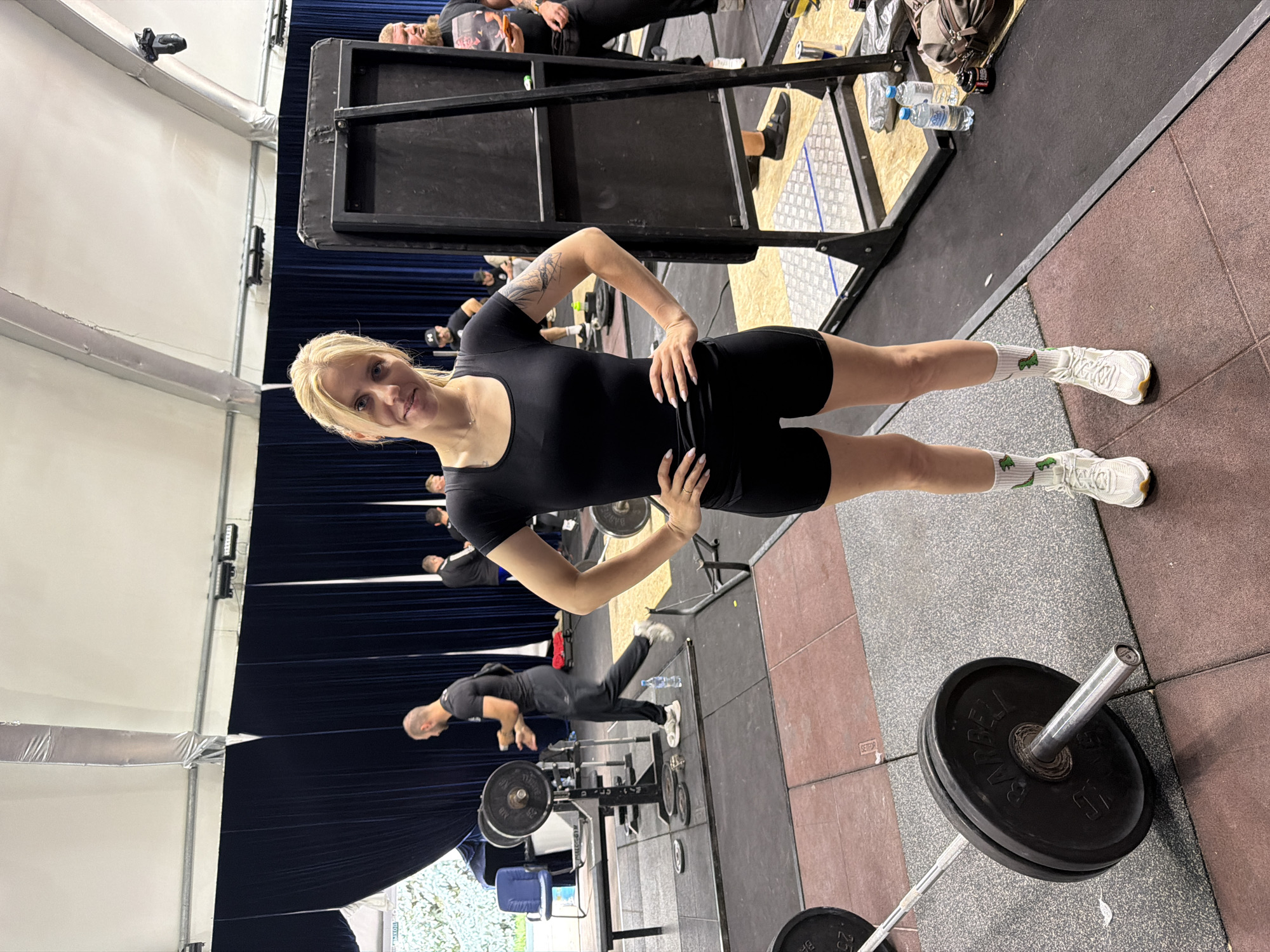 A blonde woman in a black athletic outfit stands facing the camera in a gymnasium. She has her hands on her hips and is looking at the camera with a neutral expression. She is wearing white athletic shoes with green dinosaur-patterned socks. Behind her, there are weightlifting equipment, including barbells and weight plates, and other people in athletic wear. The environment appears to be an indoor sports facility with dark blue curtains in the background and a large white tent-like ceiling structure overhead. The flooring is made of interlocking rubber mats. This scene is located in the city of Suzdal, Russia.