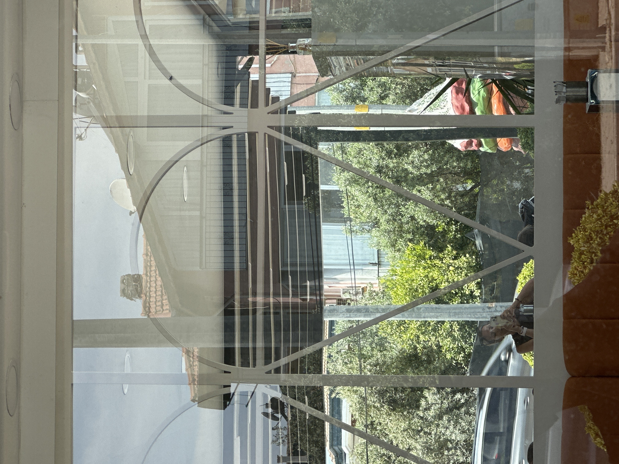The image appears to be taken from inside a building, looking out through a window with decorative metal framing. The reflection shows a building across the street with a satellite dish and what looks like blinds on the windows. Beyond that, trees and a street are visible with a person taking a photo with a smartphone on the sidewalk, next to a parked car. Laundry hanging on a line is visible in the background. Without additional context, I am unable to determine the specific location in Fethiye, Turkey.