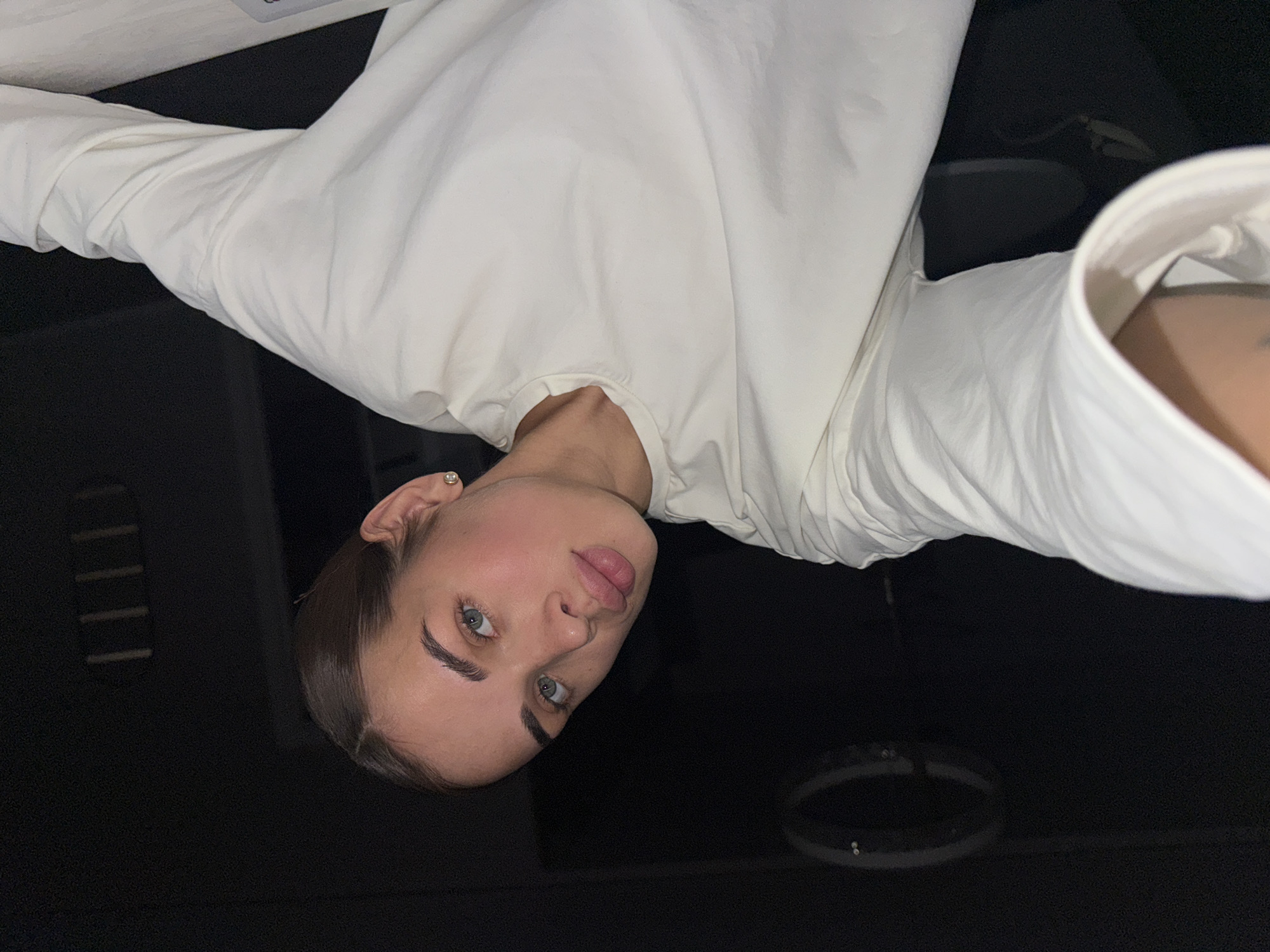 A woman with light skin, blue eyes, and dark hair pulled back from her face is oriented upside down in the frame. She is wearing a long-sleeved white top. The background is dark with some blurred, indistinct shapes that might be furniture or lighting fixtures. The image provides no information to confirm the location is in the city of Лобаново, Russia.
