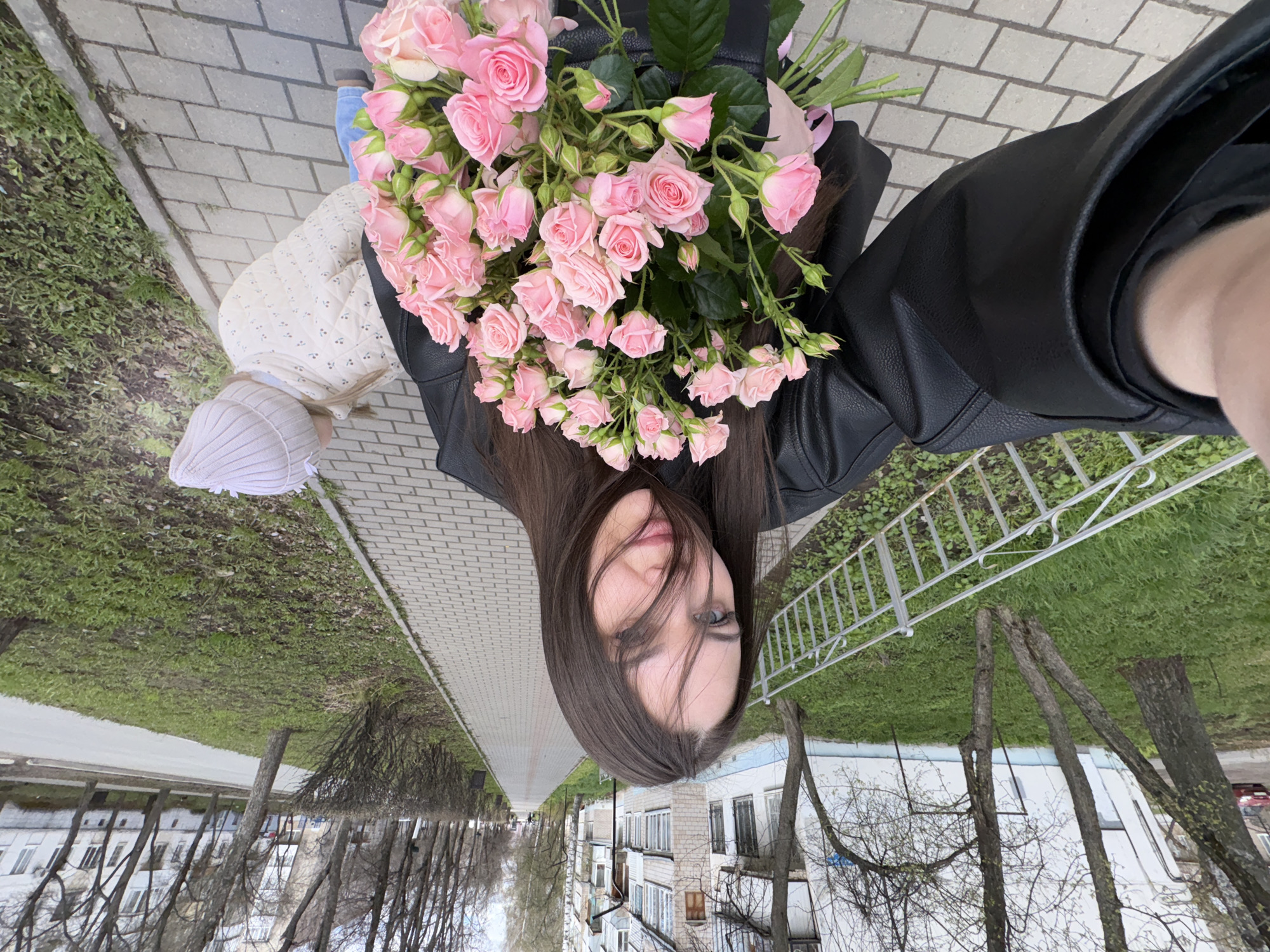 An outdoor selfie captures a young woman and a small child on a paved sidewalk. The perspective is from the woman's viewpoint, looking slightly downwards.
The primary subject is a woman with long, straight dark brown hair, wearing a black leather jacket. Her face is partially visible, with her hair falling across her eye, and she has a subtle smile. She holds a prominent, abundant bouquet of light pink spray roses. Adjacent to her, on the left side of the frame, is a small child, likely a toddler, seen from behind. The child is dressed in a cream or light beige quilted jacket and a matching ribbed beanie. A blue pant leg and a brown shoe, presumably belonging to the woman, are also visible.
They are standing on a light-colored paved sidewalk made of rectangular pavers. To their right, there is a strip of grass bordered by trees, some of which appear bare with early spring buds. In the background, a multi-story residential building constructed from light-colored brick with numerous windows is visible. To their left, another grassy area and a white metal fence with decorative elements can be seen.
The scene depicts a casual daytime outing, likely in early spring given the state of the trees. The lighting is soft and diffused, suggesting an overcast sky. The overall atmosphere is pleasant. No visible text is present in the image. The setting is Chaikovsky, Russia.