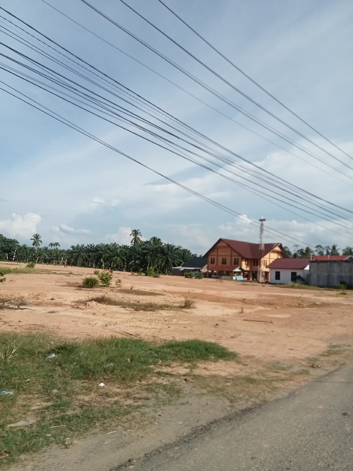 In the city of Matangpanyang, Indonesia, an open, undeveloped plot of land stretches across the frame. The ground is predominantly bare, sandy earth, with patches of sparse, dry grass and a few small bushes. In the distance, a dense line of palm trees forms a natural border, suggesting a more rural or agricultural area beyond.
Dominating the mid-ground is a cluster of buildings. The most prominent is a large, multi-story structure with a distinctive reddish-brown roof and light-colored walls, possibly yellow or beige. It appears to be a commercial or institutional building. Adjacent to it is a smaller, white building with a dark-colored roof. Further to the right, a section of a gray concrete wall is visible, possibly part of another structure. A tall, metal communication tower stands behind and slightly to the left of the main building.
Overhead, a network of electrical wires crisscrosses the sky, casting faint shadows and creating a layered effect against the pale blue sky, which is dotted with scattered white clouds. The overall impression is one of a developing area, with infrastructure in place but significant land still awaiting development. No people are clearly visible in the scene.