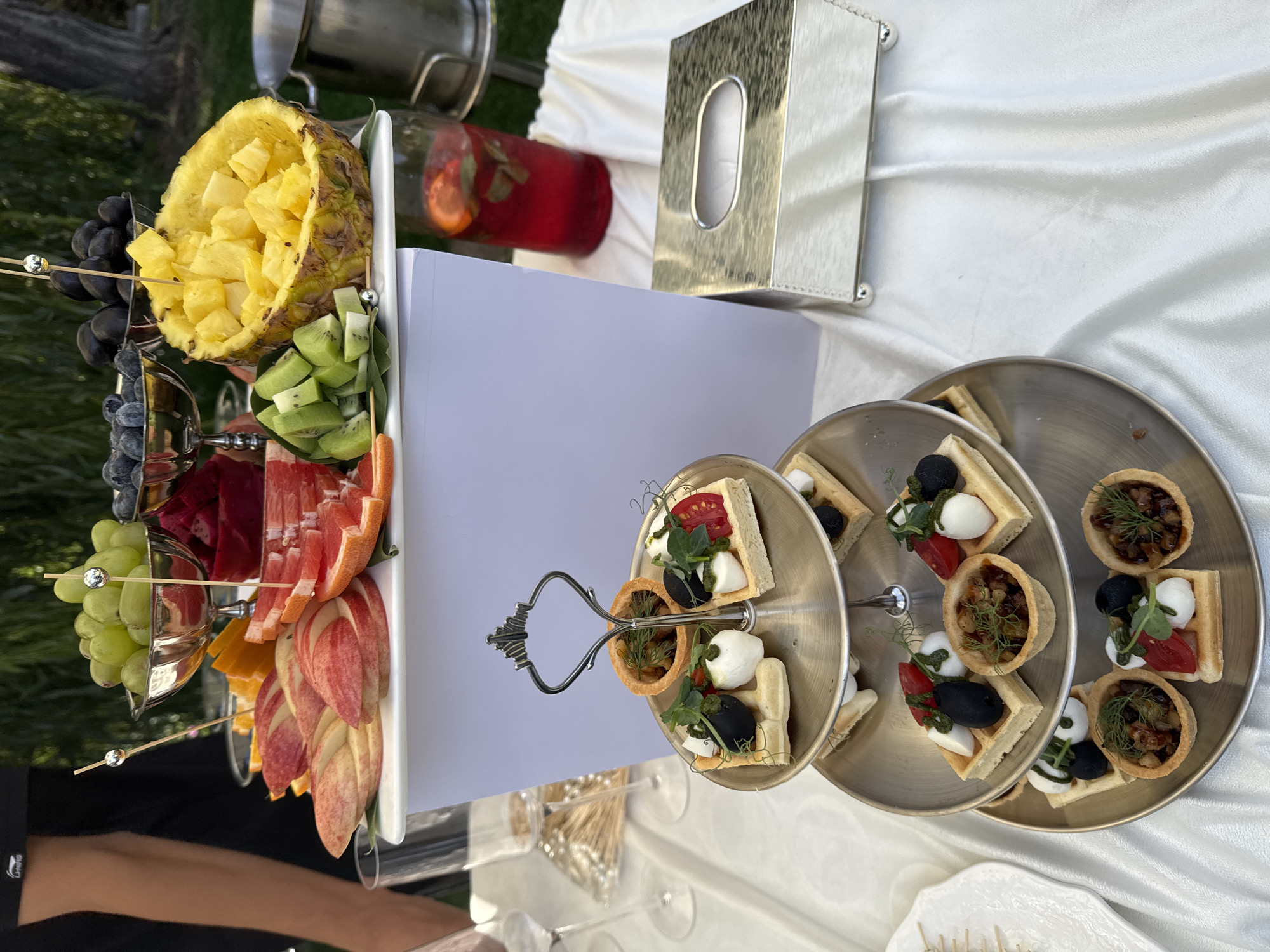 A tiered silver serving tray holds several small appetizers, each consisting of a waffle base topped with mozzarella balls, olives, chopped tomatoes, and microgreens. The top tier also has a small tartlet filled with a dark mixture. In the background, a large display of fresh fruits includes sliced apples, grapefruit, oranges, grapes, blueberries, kiwi, and diced pineapple served in a hollowed-out pineapple. The fruit display is set on a white rectangular riser. To the right of the fruit display, a tall glass pitcher contains a red beverage with mint and citrus slices. A silver tissue box sits on a white tablecloth that covers the table. A person's arm, wearing a black t-shirt, is visible on the left side of the image. The scene appears to be set outdoors, with greenery and trees visible in the background, suggesting a garden or park setting, potentially in the city of Koy Tash, Kyrgyzstan.
