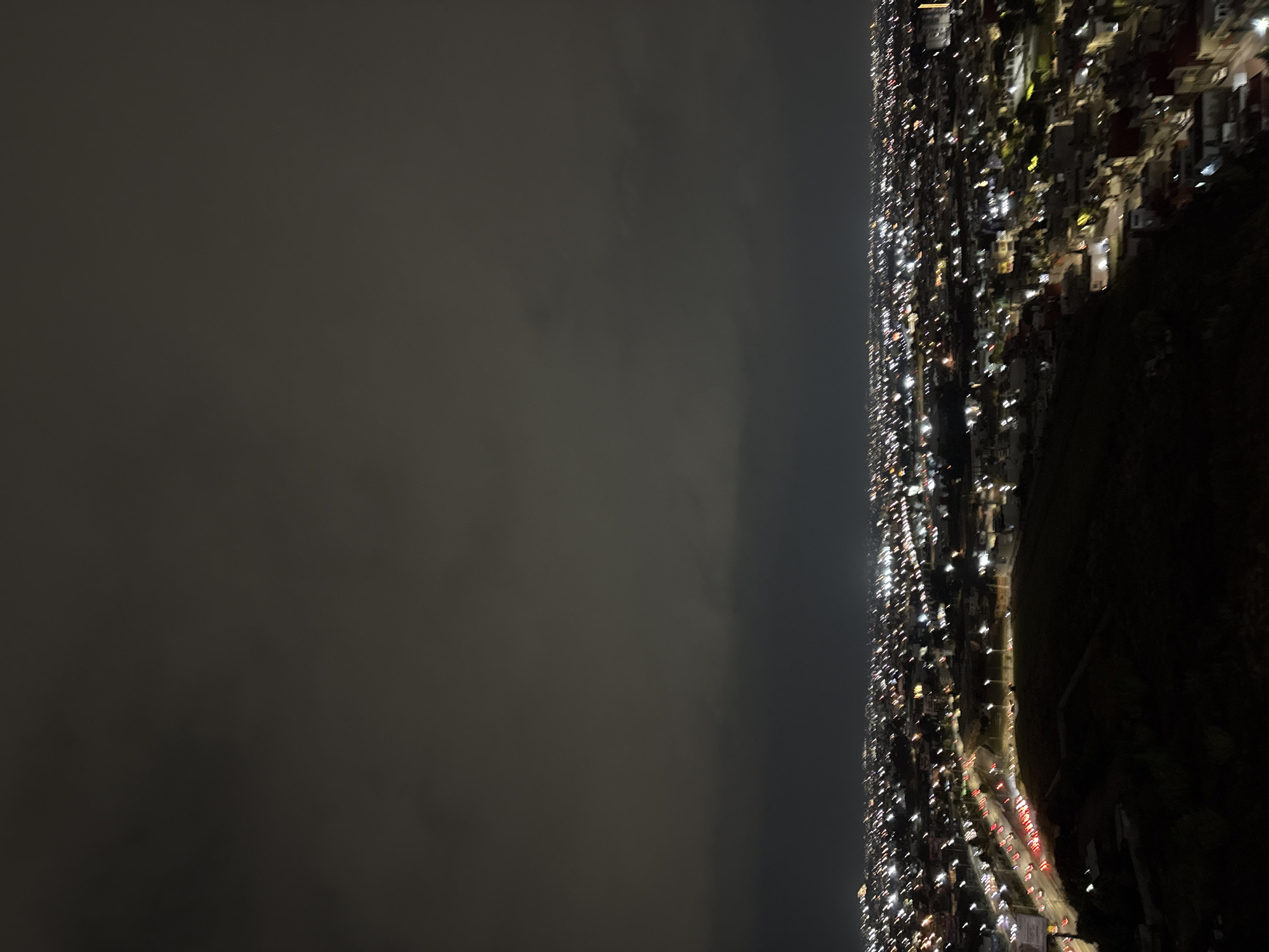 The image is an aerial view of a densely populated area in San Bernardino Tlaxcalancingo, Mexico, at night. The city lights form a bright strip on the right side of the frame. To the left, the sky is dark and obscured by what appears to be cloud cover. On the rightmost side, there is an unpopulated area. Car headlights are visible on a street. Buildings densely cover the populated area. The image is tilted.