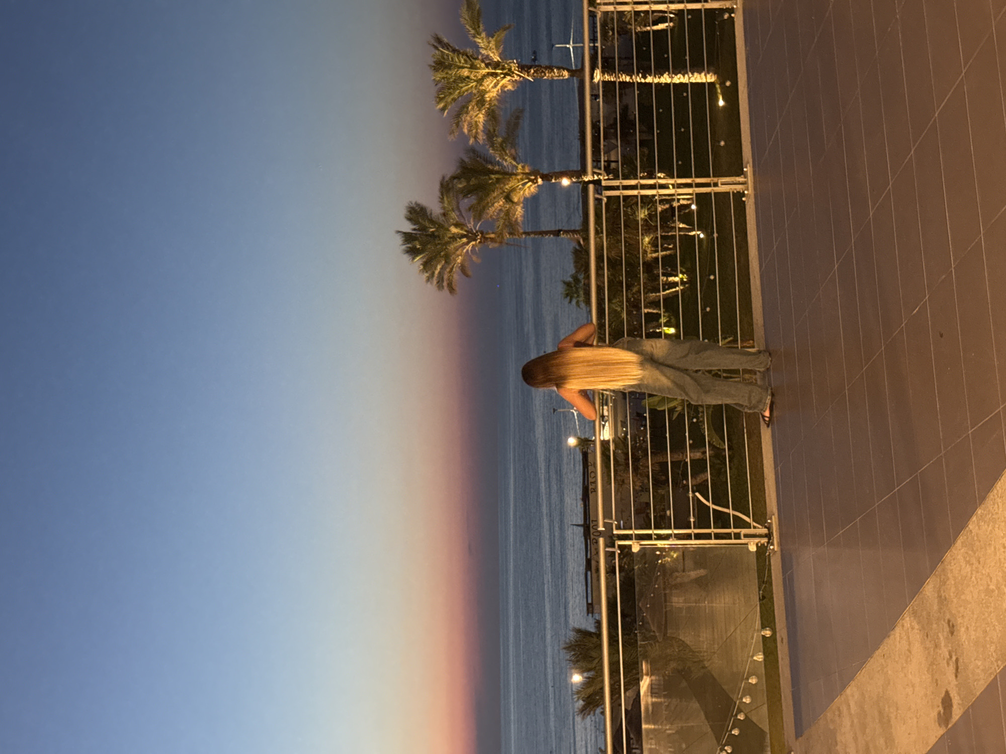 A woman stands on a tiled balcony overlooking the sea at dusk. She has long blonde hair and is wearing loose-fitting denim pants and dark sandals. She is leaning on a metal railing, facing away from the camera, towards the horizon. The sea is calm, and the sky displays a gradient of blue and pink hues from the setting sun. In the background, several palm trees are illuminated, casting shadows. Lights are strung along the railing and are also visible on a lower level of the structure, suggesting an outdoor dining or recreational area. The tiled floor of the balcony is a warm brown color. The scene appears to be a scenic overlook or terrace, possibly at a resort or restaurant, in Isola delle Femmine, Italy, given the coastal setting and palm trees.