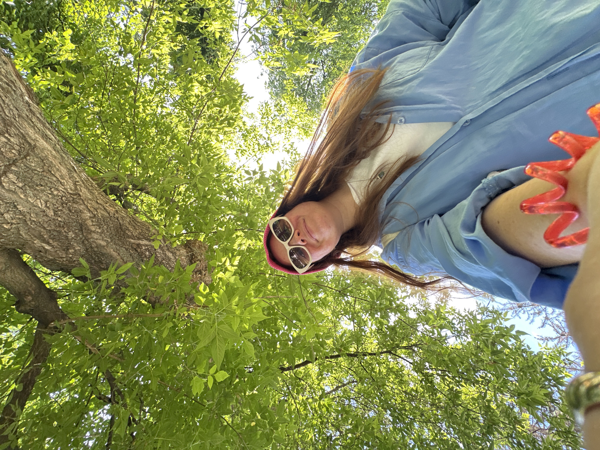 A low-angle, "worm's-eye view" photograph captures a person looking down towards the camera from beneath a dense, vibrant green tree canopy. The subject, positioned in the lower part of the frame, has long brown hair and wears a light blue long-sleeved shirt over a white top. White-framed sunglasses are perched on their head, slightly obscuring their eyes, atop a pink or red beanie. A distinctive bright red, spiky or zig-zagged bracelet adorns their right wrist, and a metallic ring is visible on a finger.
The background is dominated by the lush foliage of a large tree, with bright sunlight filtering through the leaves, creating a dappled effect and indicating a sunny day. A significant portion of a tree trunk with rough, textured brown bark is visible on the left side of the frame, extending upwards. Patches of clear blue sky are visible through the gaps in the leaves.
The image depicts a relaxed outdoor portrait taken in a natural setting, likely a park or garden, in Nizhny Novgorod, Russia. The unusual perspective highlights the height of the tree and the open sky above. There is no visible text in the image.