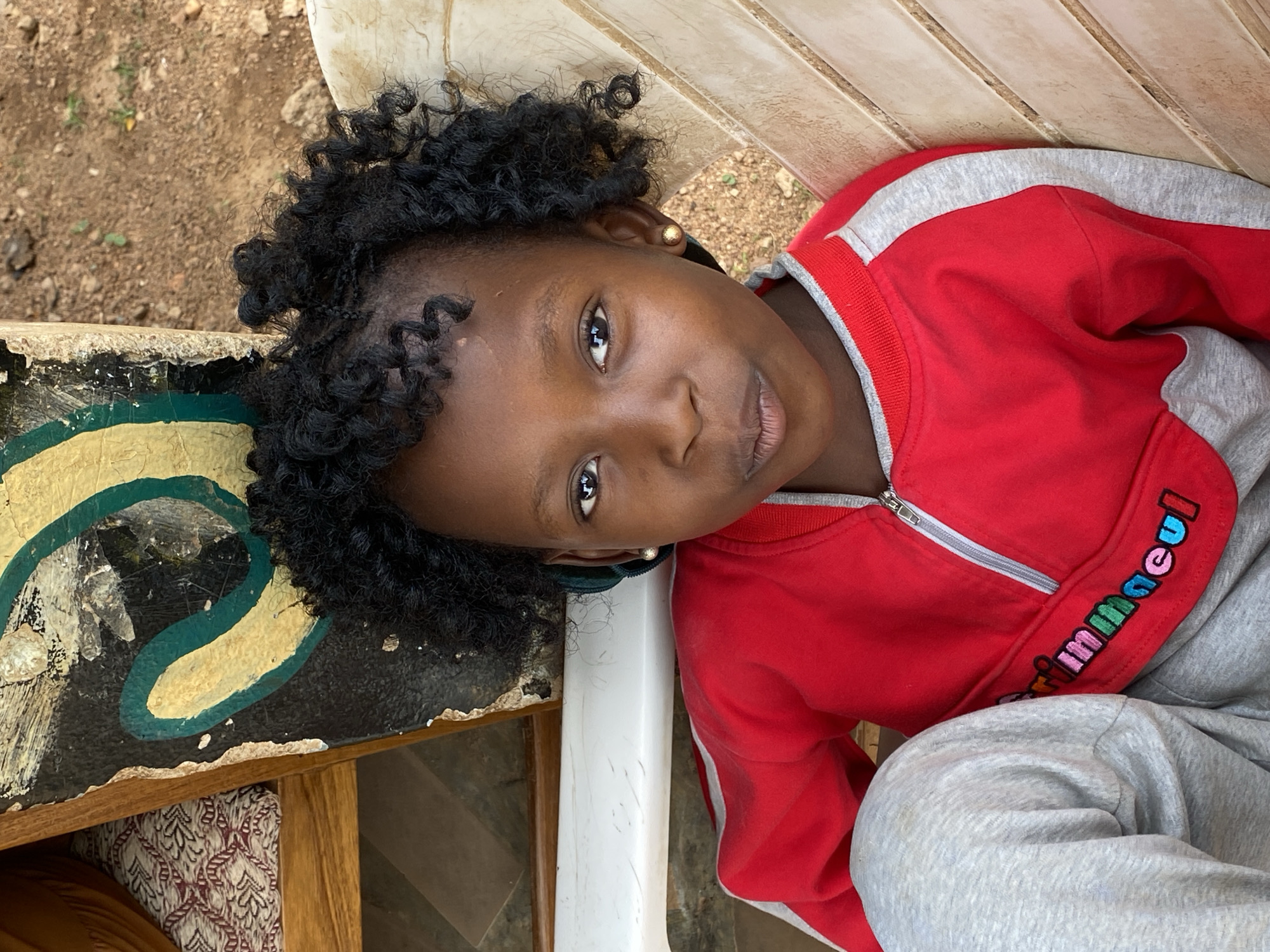 A young girl with curly black hair is seated on a white plastic chair. She is wearing a red and gray tracksuit with a zipper and embroidery. She is looking at the camera. The background is a mix of natural elements and man-made objects, including a painted table and some soil. While the city is mentioned, I cannot verify the location without further information visible in the image.