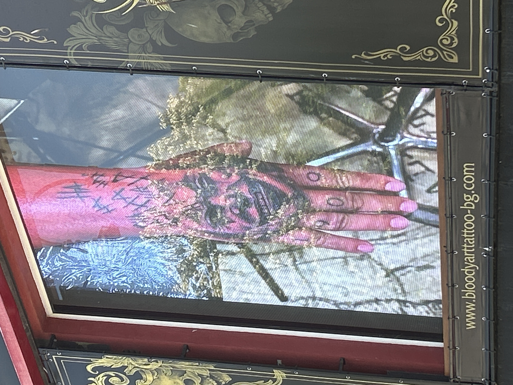 The image shows a close-up of a screen displaying a tattoo. The tattoo appears to be on a hand, with a stylized face depicted on the palm and "HA HA HA" written on the back of the hand. The hand is pink, and the tattoo ink is black and red. There are also some leafy elements in the image, possibly representing foliage. To the left and right of the screen are dark panels with gold decorative elements and a skull motif. Below the screen, a dark banner with white text reads "www.bloodyarttattoo-bg.com". The overall context suggests this is a display at a tattoo parlor or event, likely in Varna, Bulgaria, given the ".bg" domain name. The screen itself is framed by a dark bezel and mounted on a red structure.