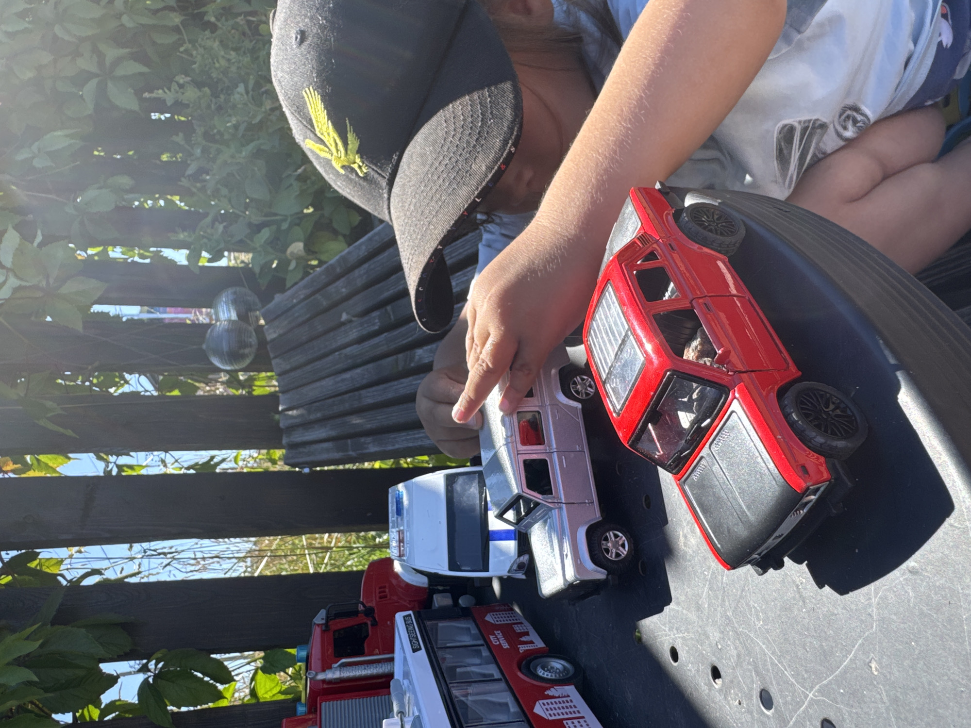 In Kempelevo, Russia, on a sunny day, a child wearing a black baseball cap with a yellow eagle emblem plays with toy vehicles outdoors. The child's hand is visible, reaching towards a red toy car. The toy vehicles include a red fire truck, a white bus with "CITY SERVICE" branding, a silver SUV, a white and blue van, and a red sedan. The toys are arranged on a dark, textured surface, possibly a table or patio. Behind the toys, a wooden fence and lush green foliage are visible, suggesting a backyard setting. The sunlight casts shadows across the scene.