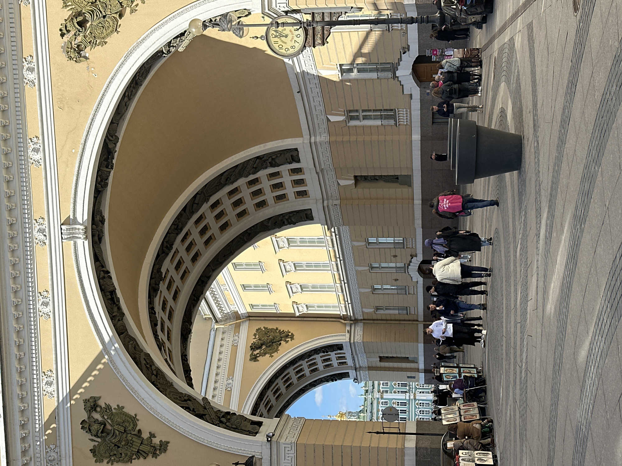 The image depicts a grand architectural scene in Saint Petersburg, Russia, dominated by a large, ornate archway on the left, likely part of a historic building complex such as the General Staff Building. The archway is lavishly decorated with white classical moldings, sculptures, and gold accents, leading into a spacious public square.
Through the arch, a sunny urban landscape unfolds. On the right, a multi-story building with numerous windows and classical elements, painted in a light yellow hue, is visible. Further in the distance, another distinct light-blue building with a golden dome can be seen against a clear blue sky, hinting at the presence of the Winter Palace or a similar landmark.
The scene is bustling with people enjoying a bright, sunny day. Pedestrians are seen strolling, standing in groups, and observing their surroundings. A significant activity involves street artists who have set up easels and displays along the paved square, showcasing various paintings, including portraits and landscapes, to potential customers. Large, dark, decorative planters are arranged on the patterned grey and beige paved ground.
A prominent detail is a vintage-style street clock hanging from the archway, clearly showing the time as approximately 1:20. The clock face also features the text "ВАЛДАЙ" (Valday). One person in the foreground is wearing a bright pink top with partially visible white text that appears to read "CITY..." followed by another word. The clear shadows suggest bright sunshine, indicating a daytime setting. The overall ambiance is that of a lively historical city square.